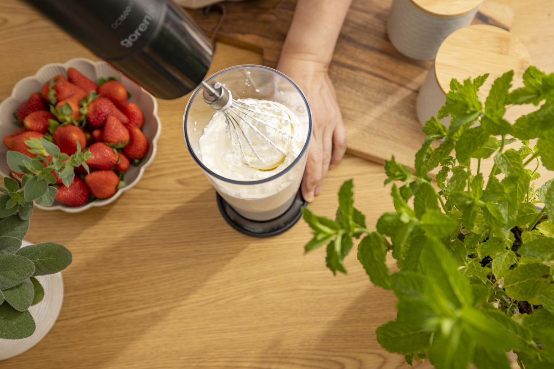 Blender ręczny ubija śmietanę w szklanym pojemniku, obok truskawek i mięty.