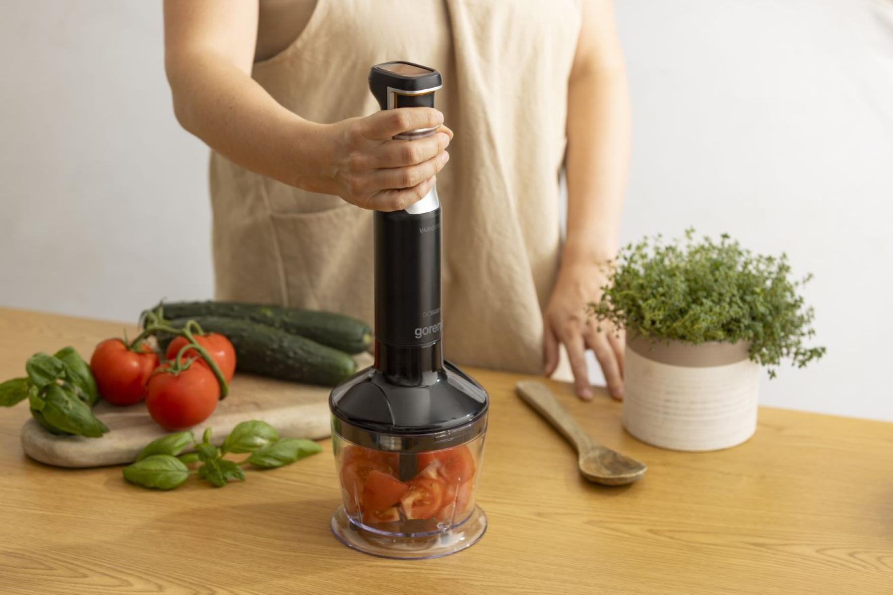 Ręka trzyma czarny blender ręczny nad pojemnikiem z posiekanymi pomidorami.
