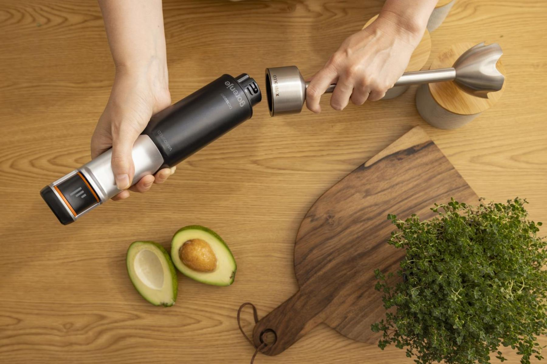 Ręce składają czarny blender Gorenje na drewnianym stole z awokado i ziołami.