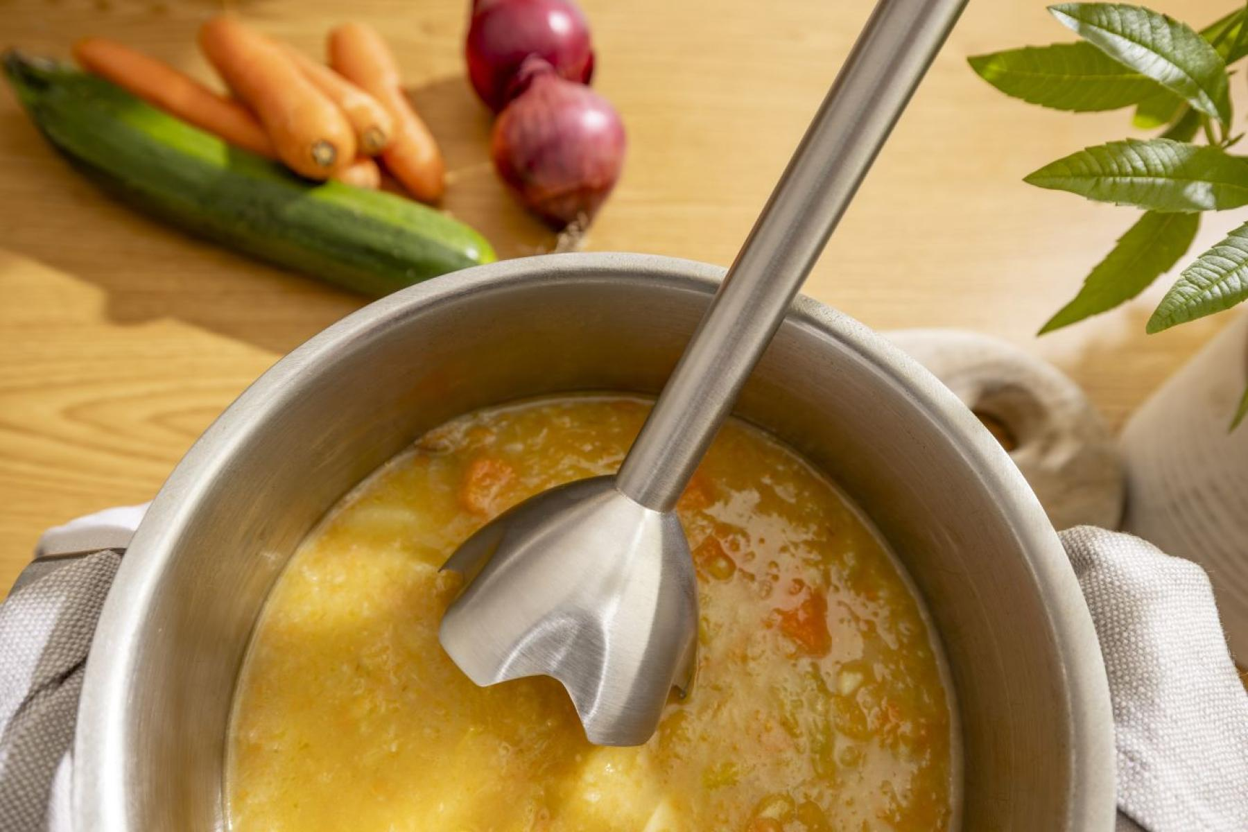 Blender ręczny w garnku z żółtą zupą, obok marchewki, cukinia i cebula.