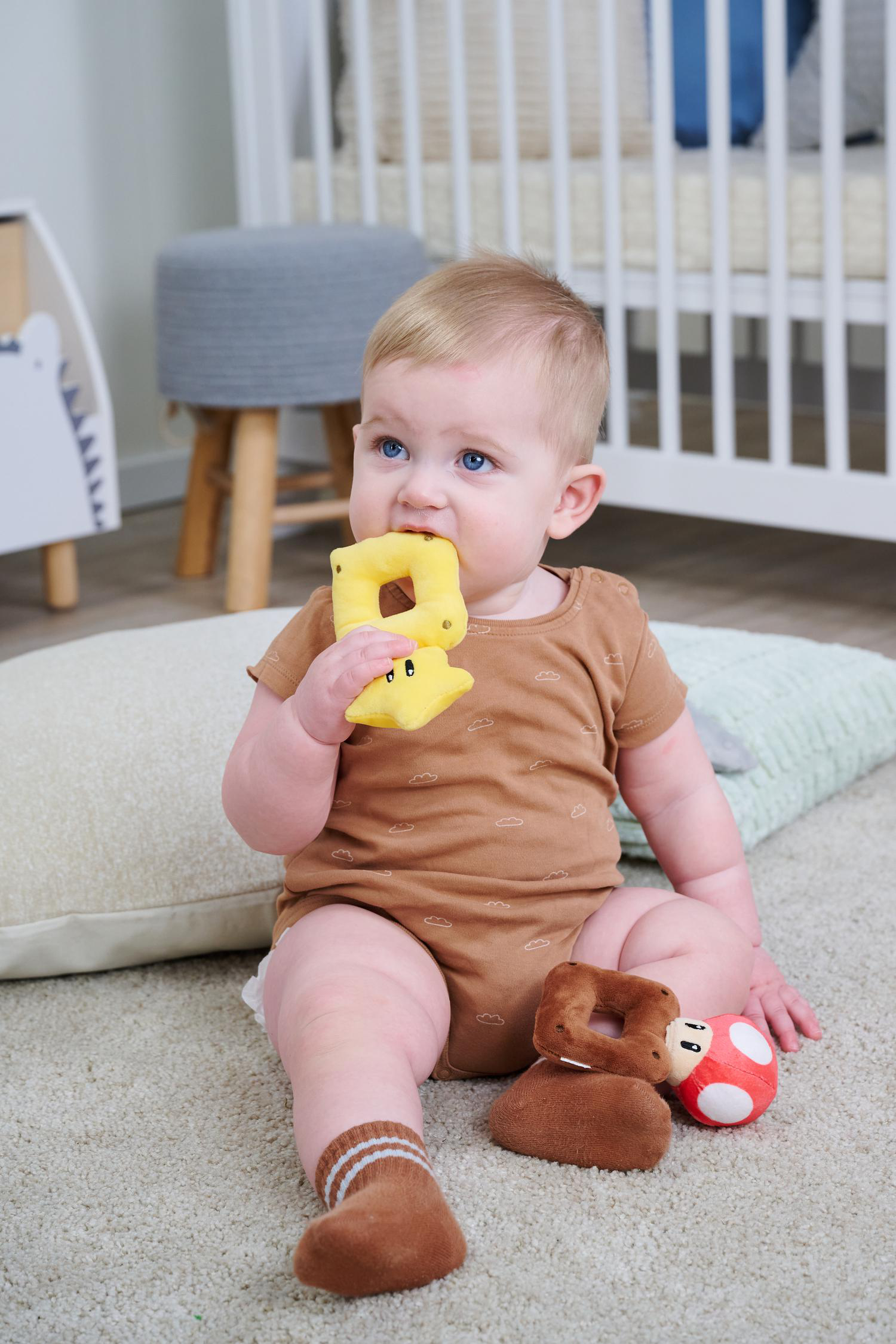 Ein Baby sitzt auf dem Boden und kaut auf einem gelben Spielzeug. Ein Pilzspielzeug liegt daneben.