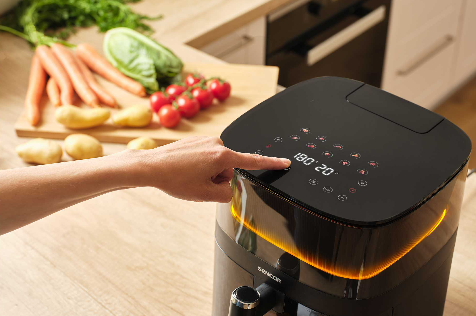 Egy kéz digitális beállításokkal működtet egy fekete air fryer-t, zöldségekkel a háttérben.