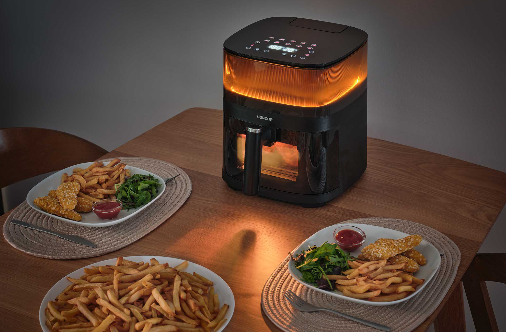 Egy fekete légkeveréses fritő, narancssárgán világító felsőrésszel, faasztalon, két tányér sültkrumplival.