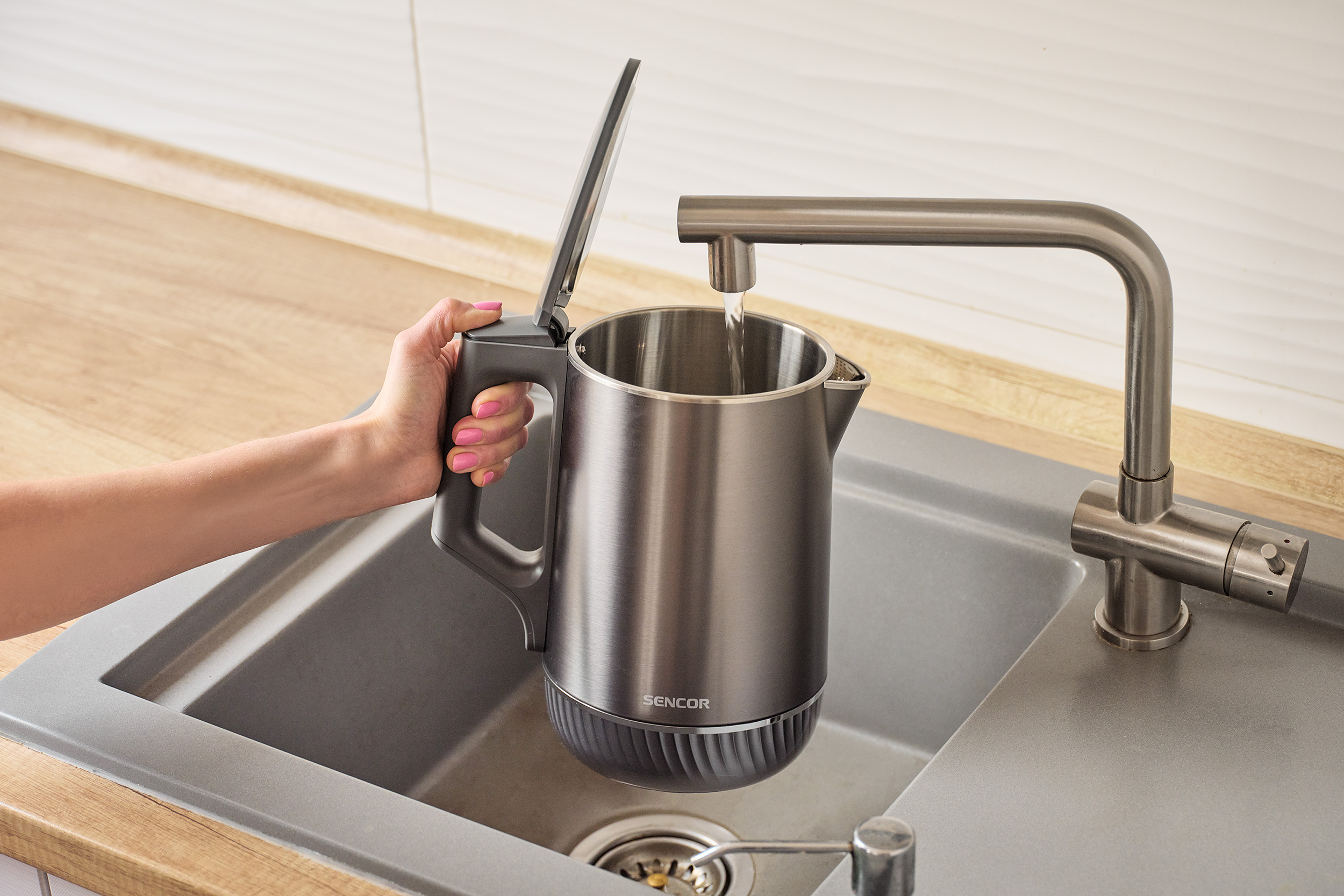 Egy kéz tölt egy szürke vízforralót egy csap alatt egy mosogatóban.