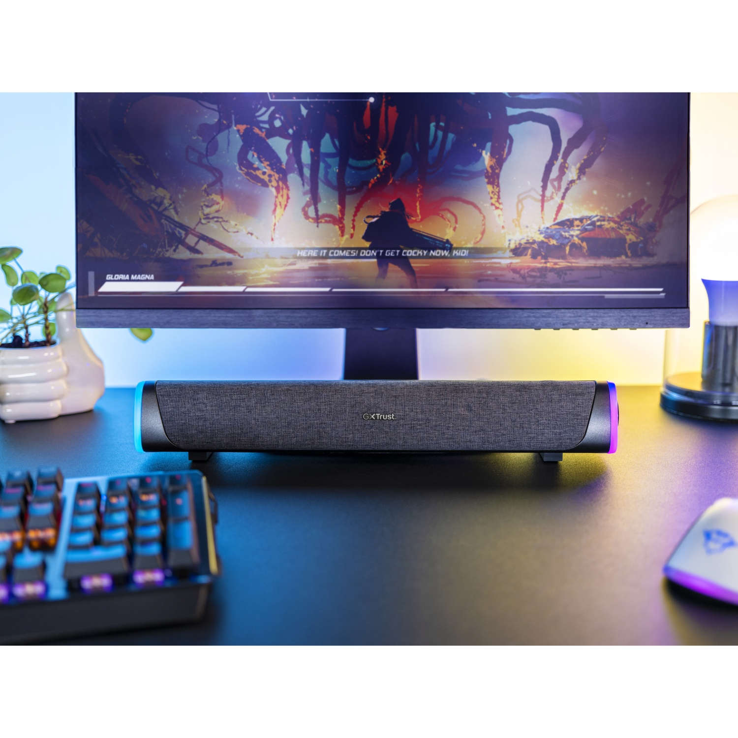 A dark gray soundbar with RGB lighting sits on a desk below a monitor displaying a video game.