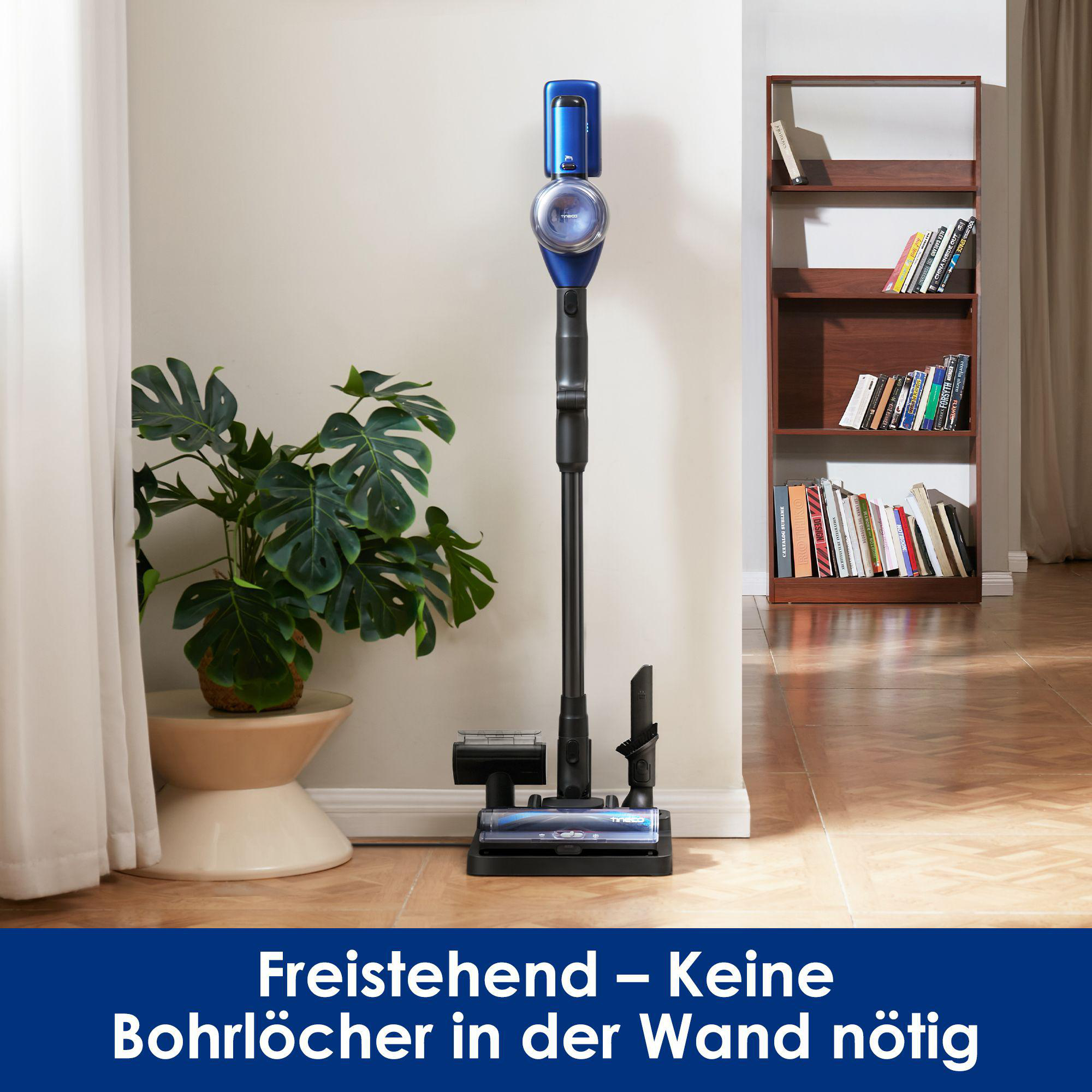 Blauer Akku-Staubsauger steht auf Ladestation neben Pflanze und Bücherregal.