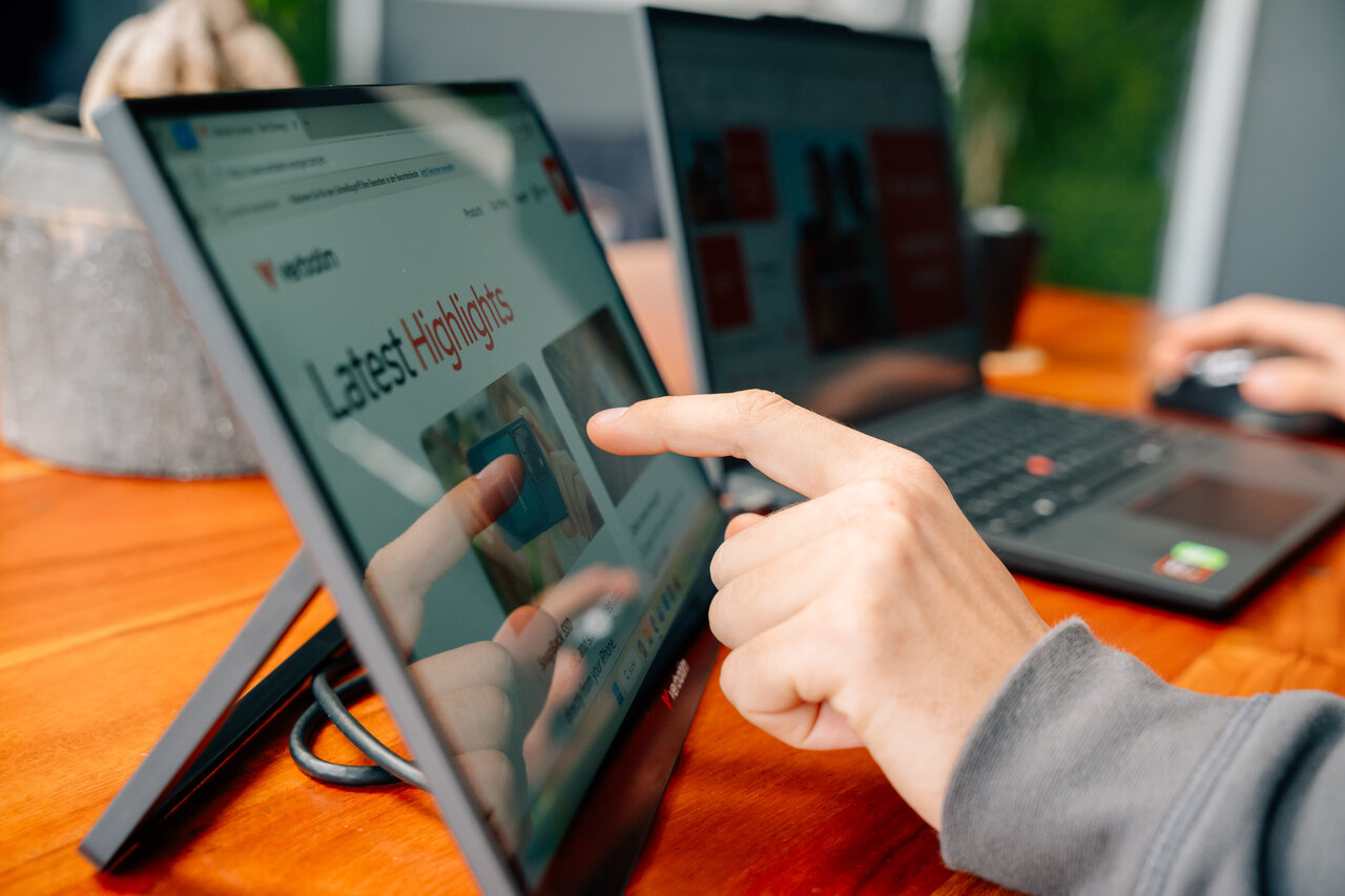 Le mani di una persona usano un laptop e un monitor portatile su una scrivania di legno.