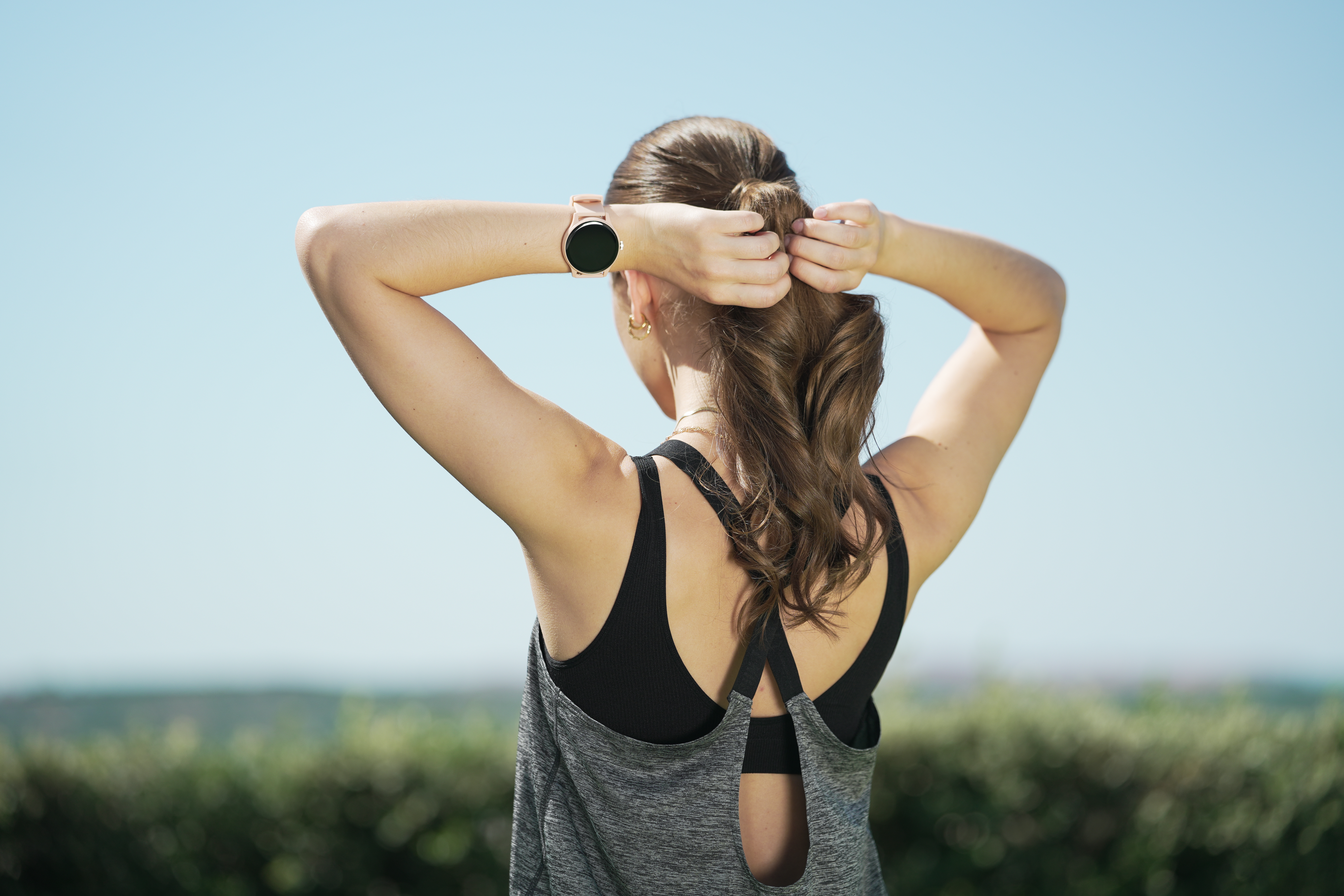 Una donna di spalle, con uno smartwatch, che si lega i capelli castani in una coda di cavallo contro un cielo azzurro.