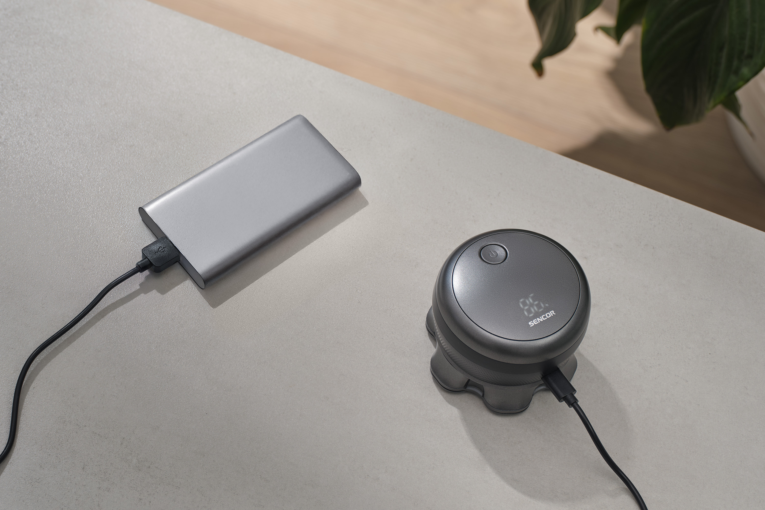 Szürke powerbank és SENCOR eszköz kábelekkel az asztalon. Növény a háttérben.