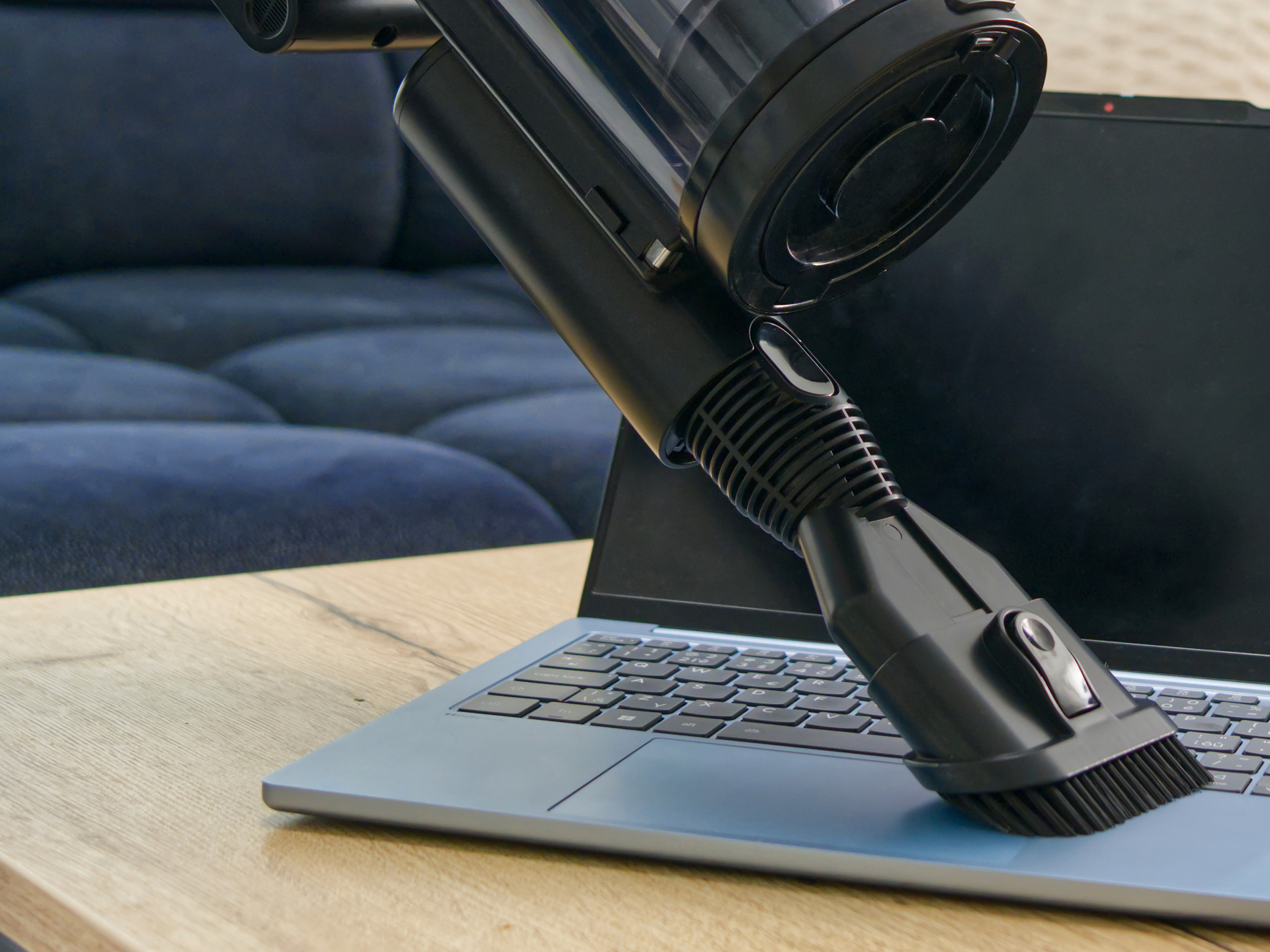 Egy fekete kézi porszívó tisztít egy kék laptop billentyűzetét egy faasztalon.