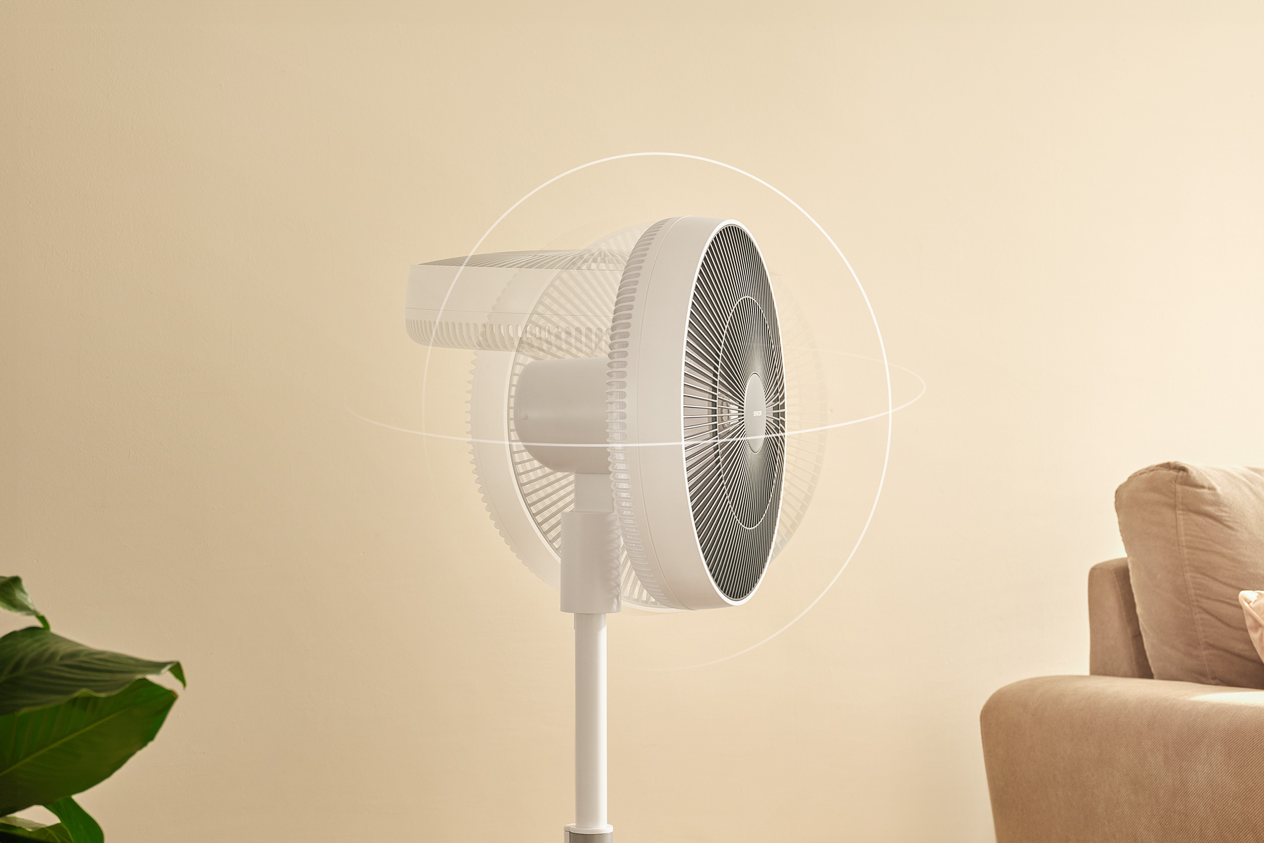 Egy fehér álló ventilátor lengéscsillapít egy krémszínű fal előtt, kanapéval a háttérben.