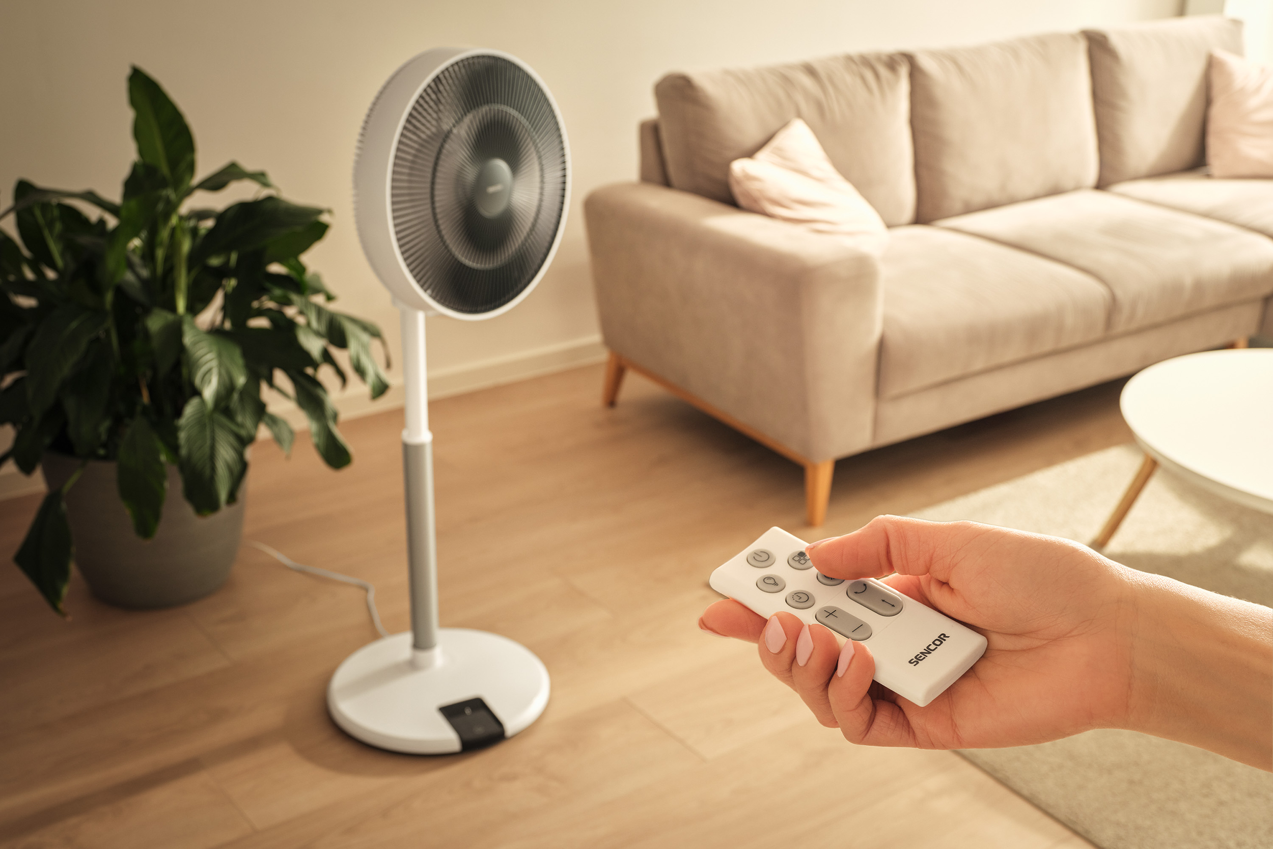 Egy kéz távirányítót tart egy ventilátor, kanapé és növény előtt.