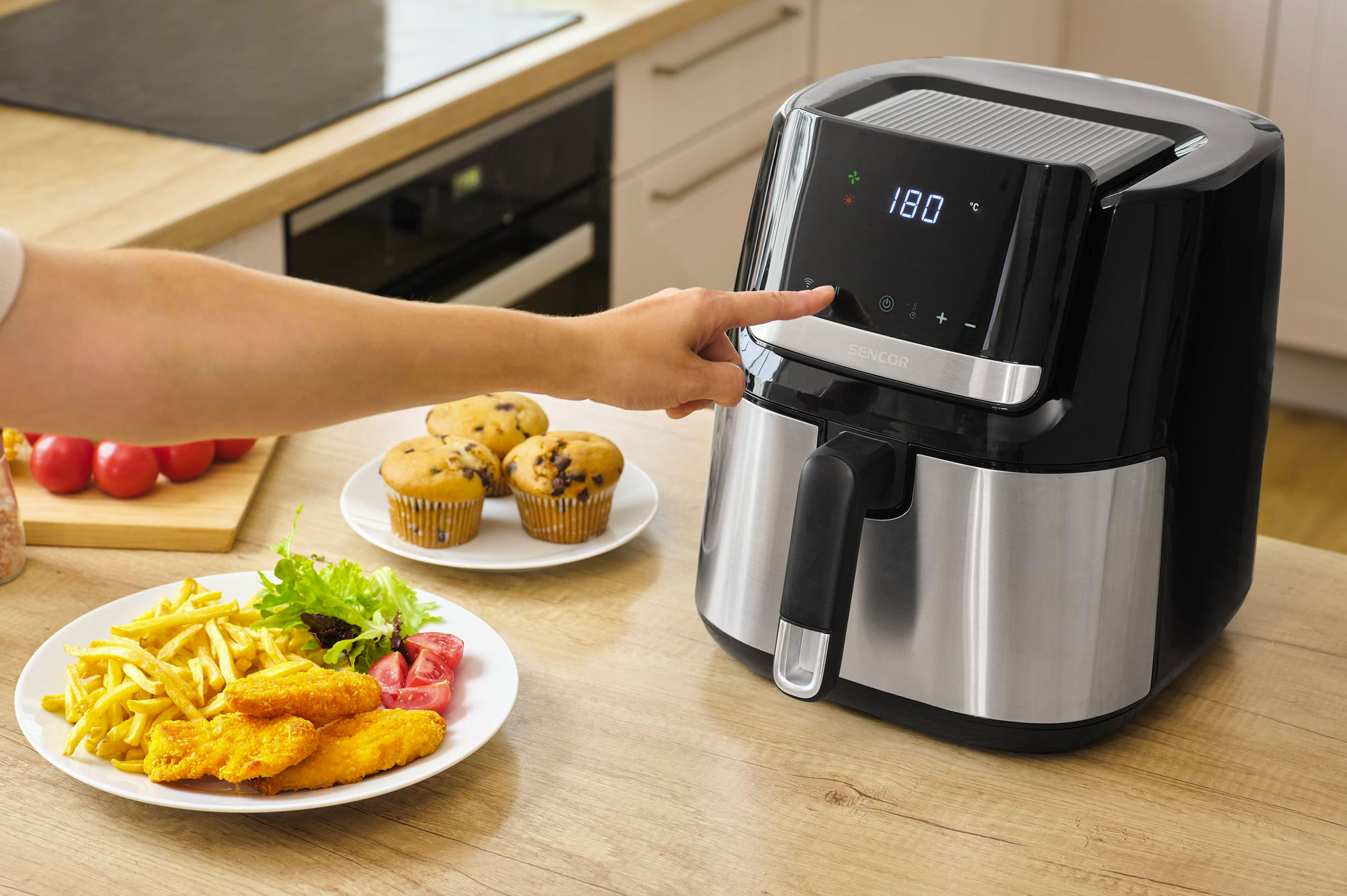 Egy kéz fekete, digitális kijelzős légkeveréses fritőzöt kezel. Mellette muffint és sült krumplis, rántott falatkákat tartalmazó tányért látni.