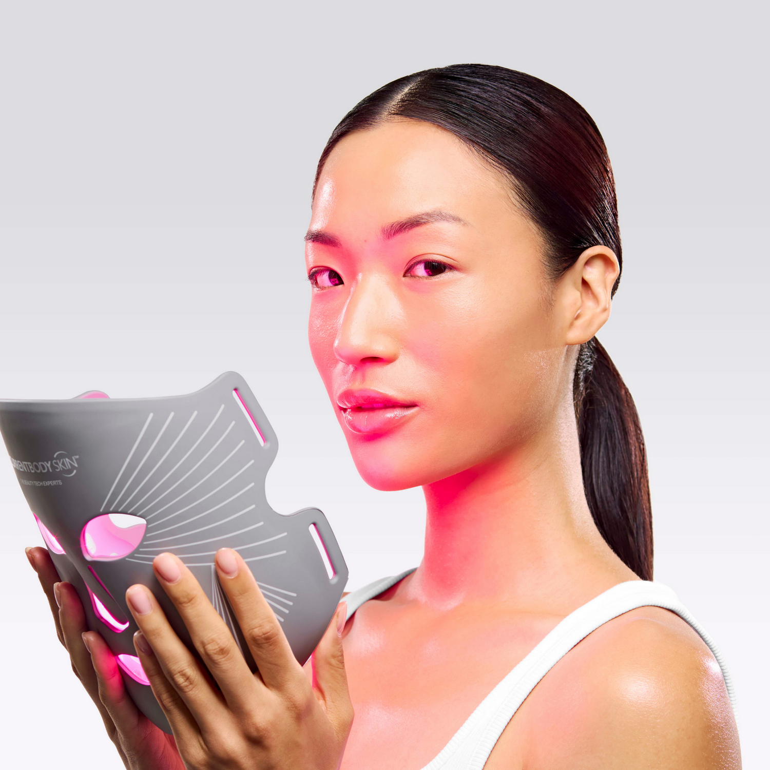 Una mujer sostiene una mascarilla facial LED gris con luces rosas frente a su rostro.