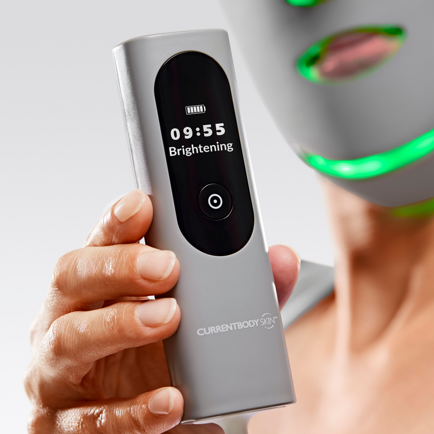 Una mano sostiene un dispositivo gris con pantalla. Una mascarilla con luces verdes al fondo.