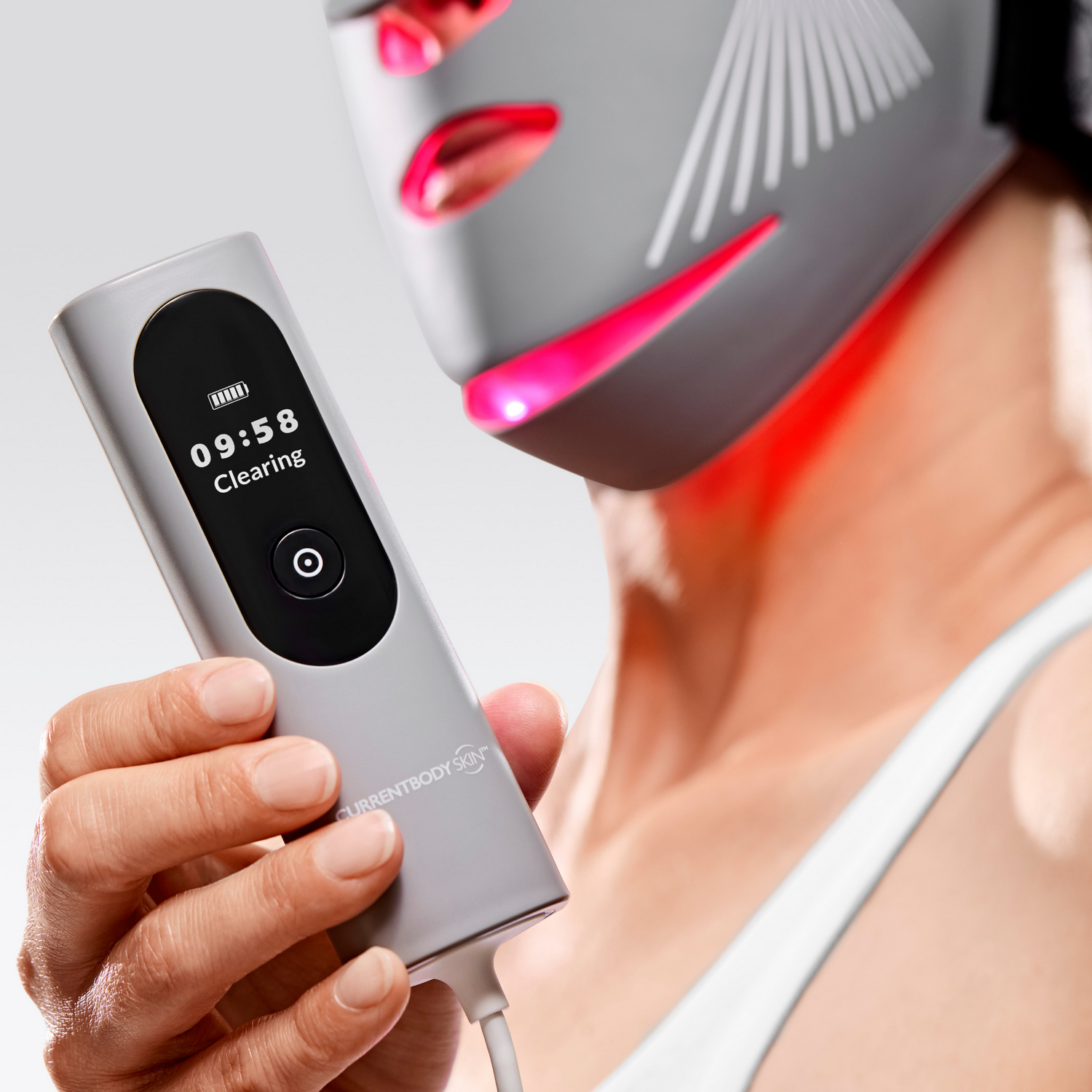 Una mano sostiene un dispositivo gris con pantalla. Una mascarilla con luces rojas al fondo.