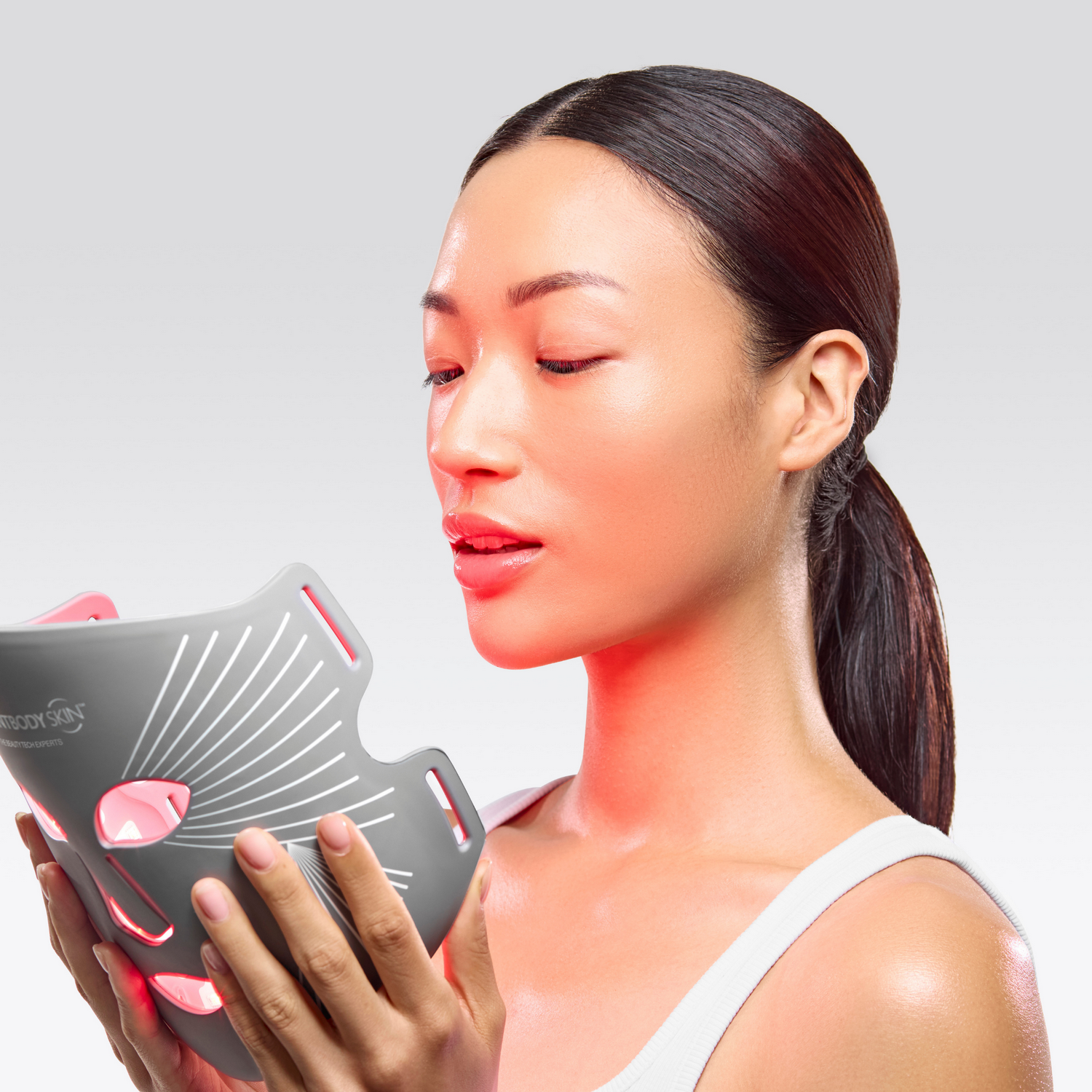 Mujer sosteniendo una mascarilla LED gris con luces rojas frente a su rostro.