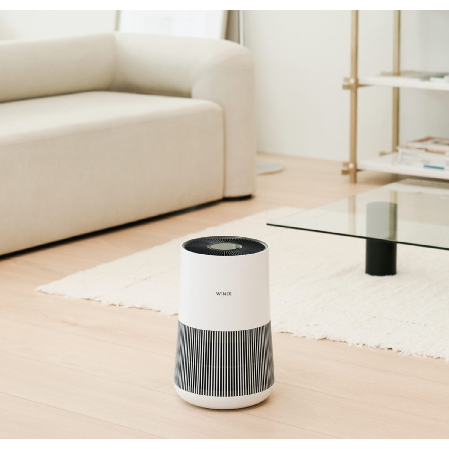 A white Winix air purifier sits on a wooden floor next to a cream sofa and rug.