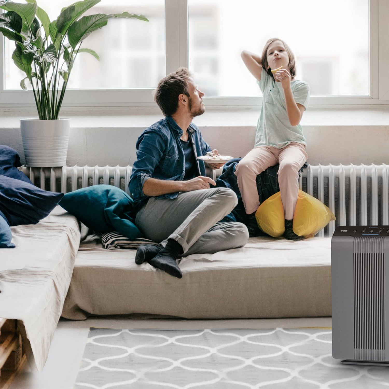 Man and child sit on couch by window; air purifier in foreground.