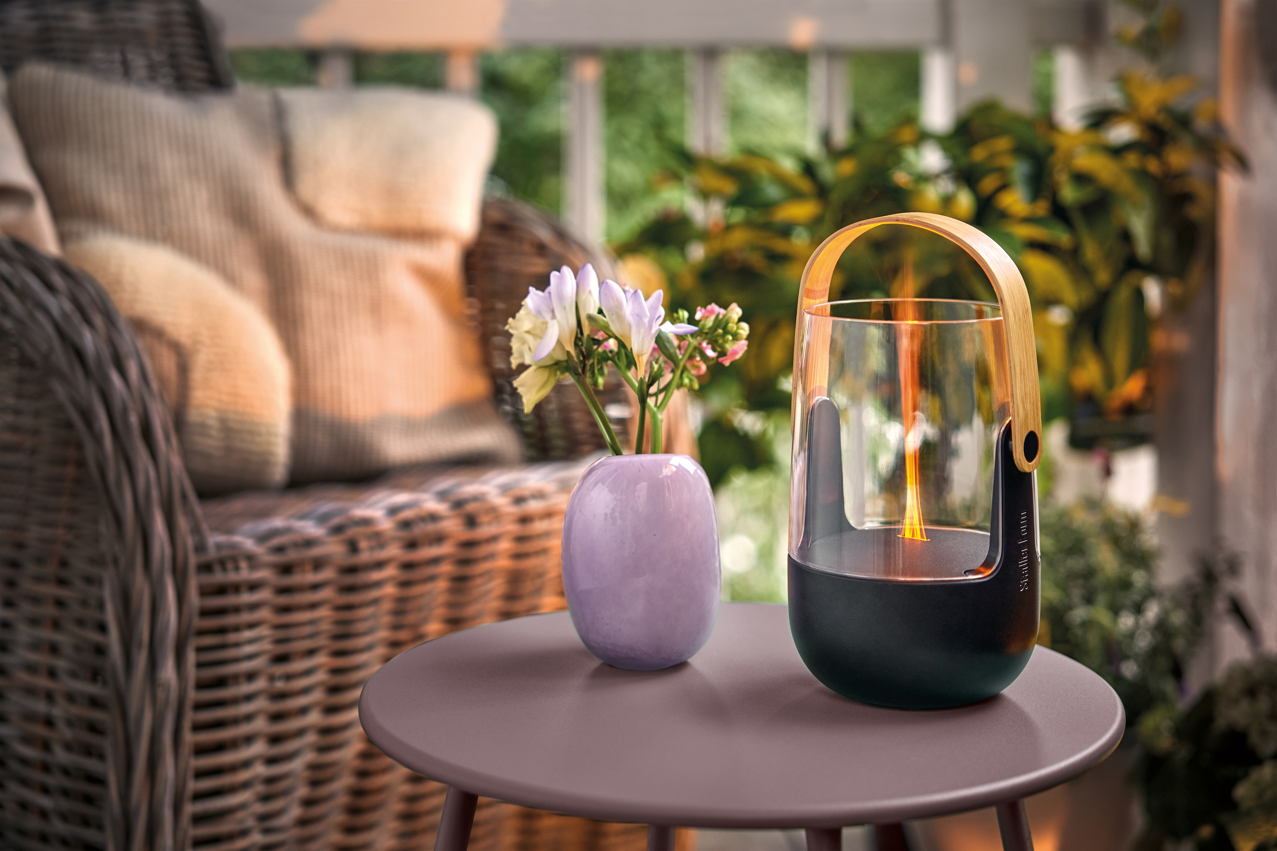 Ein Korbstuhl mit Kissen, ein lila Vase mit Blumen und eine Laterne mit Flamme auf einem Tisch.
