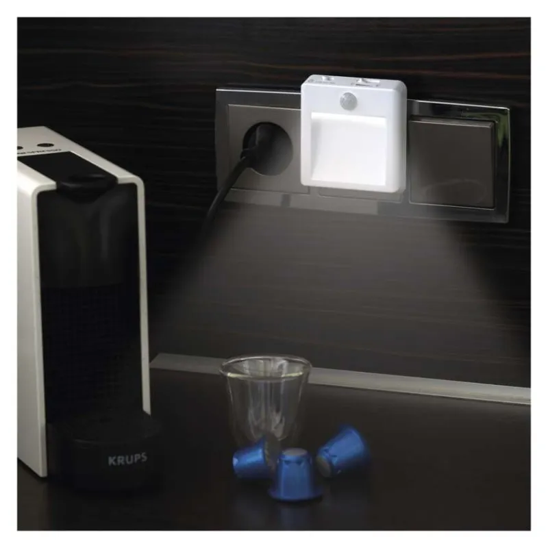 Egy fehér LED-es éjszakai lámpa adapter egy konnektorban, kávéfőző mellett.