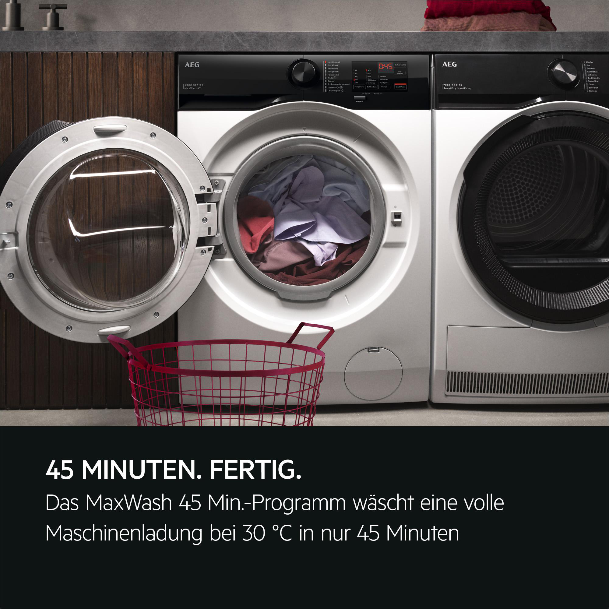 Eine offene Waschmaschine mit Wäsche und daneben ein Trockner. Ein roter Korb steht davor.