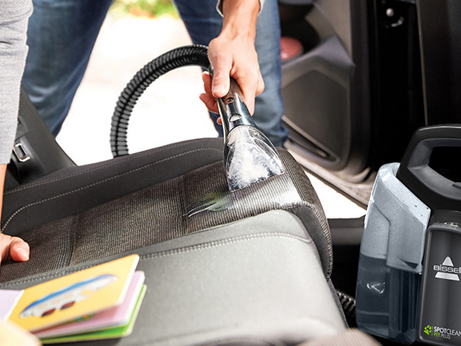 Limpieza de un asiento de coche con un dispositivo de mano y manguera. Un libro descansa cerca.