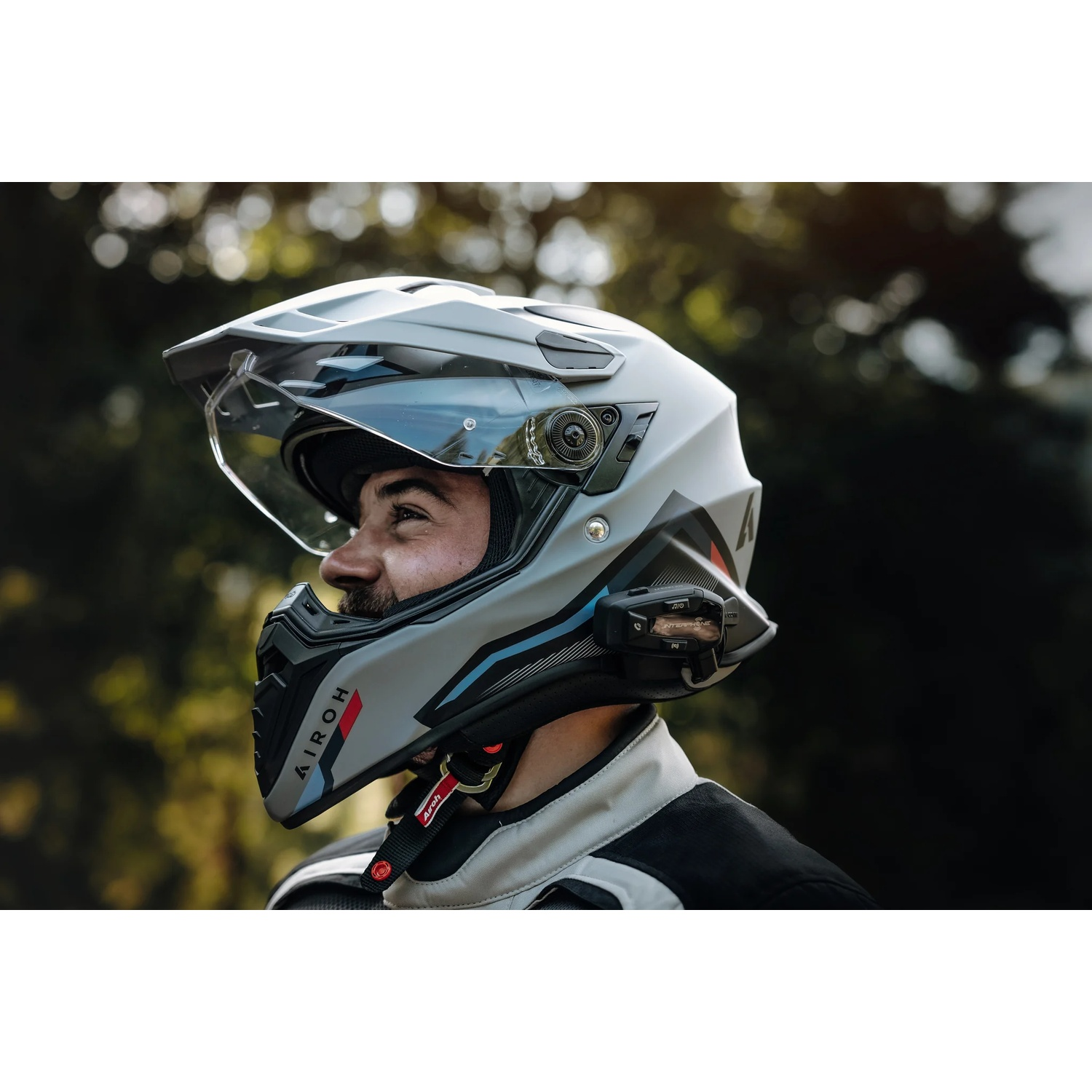 Uomo con casco da moto grigio con visiera e dispositivo di comunicazione, sfondo sfocato.