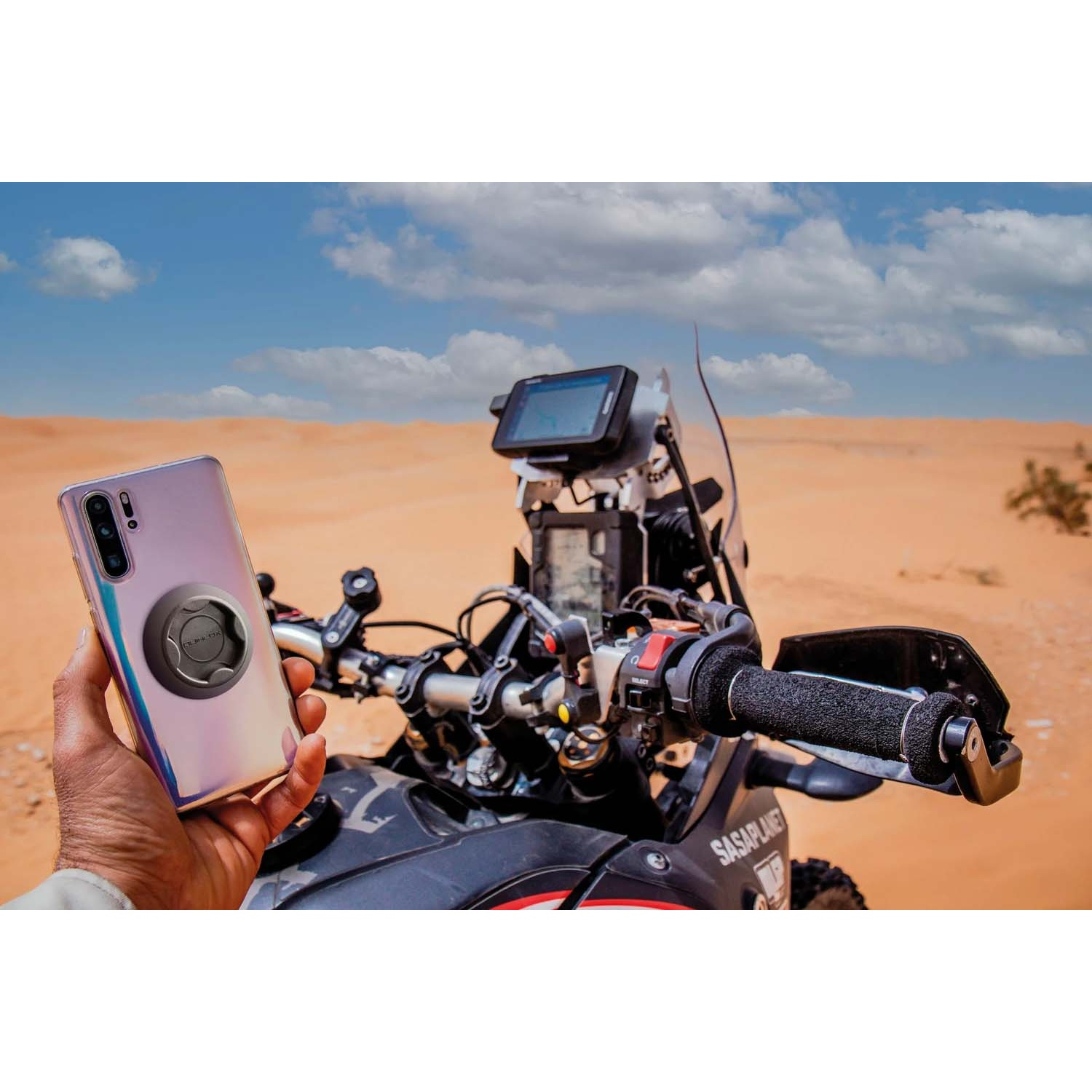 Mano tiene telefono davanti a moto nel deserto sotto cielo blu.