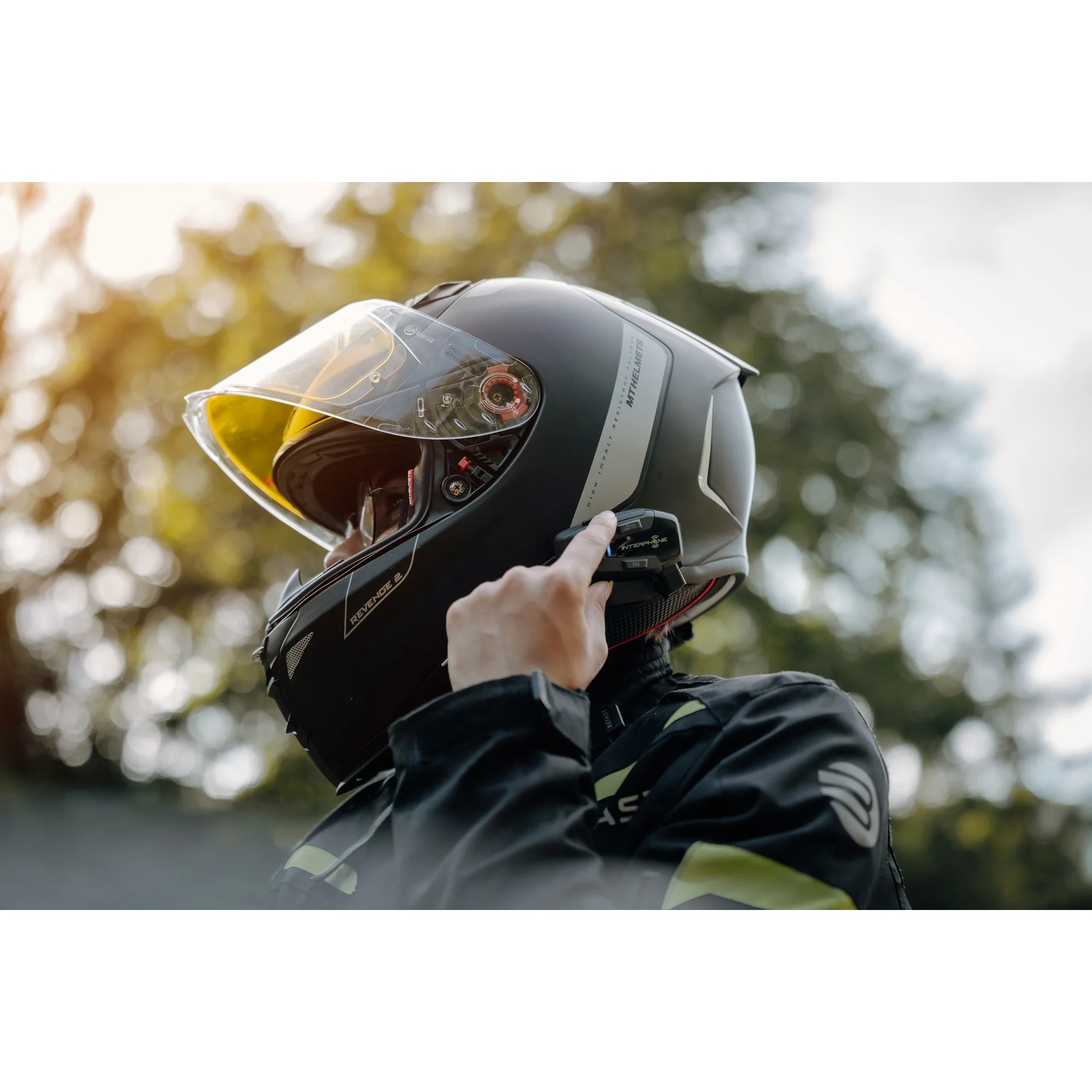 Uomo con casco da moto nero, visiera gialla, che usa un interfono.