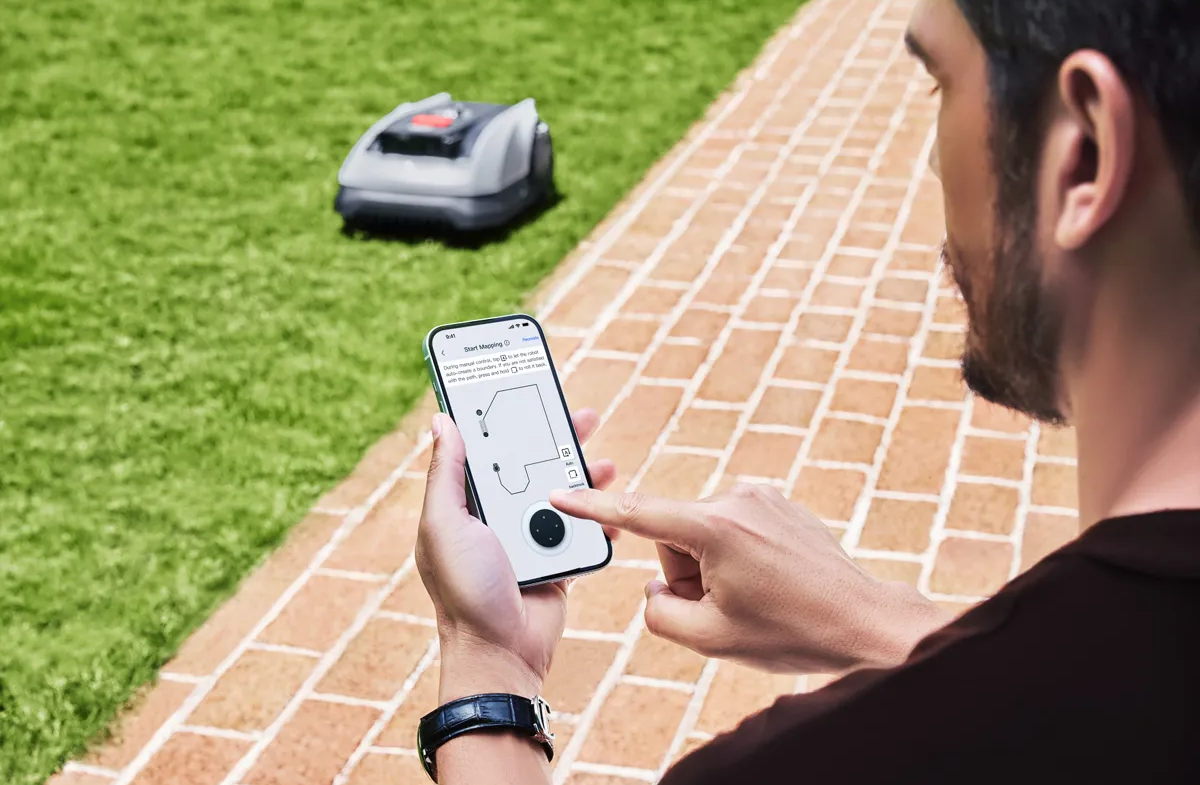 Mann bedient Mähroboter per Smartphone auf gepflastertem Weg neben Rasen.