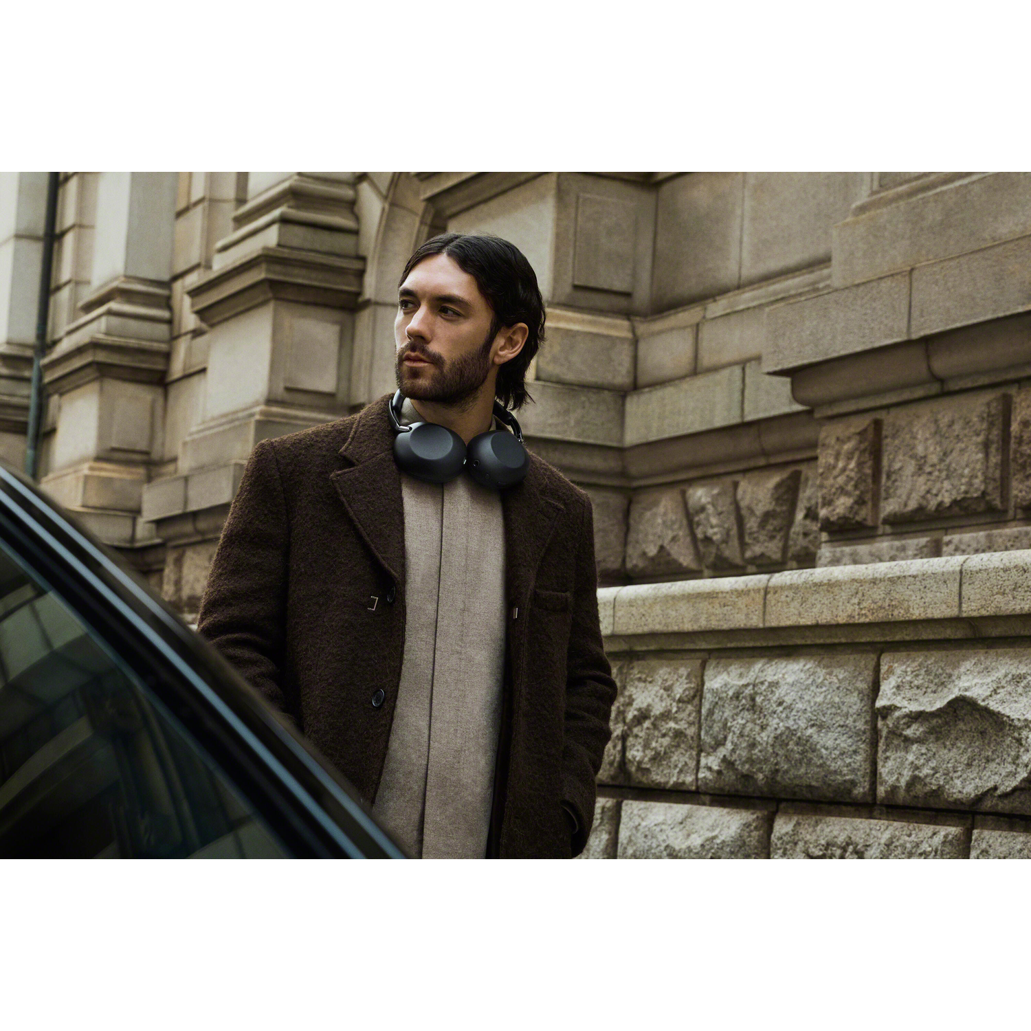 A man wearing headphones stands in front of a stone wall next to a car.