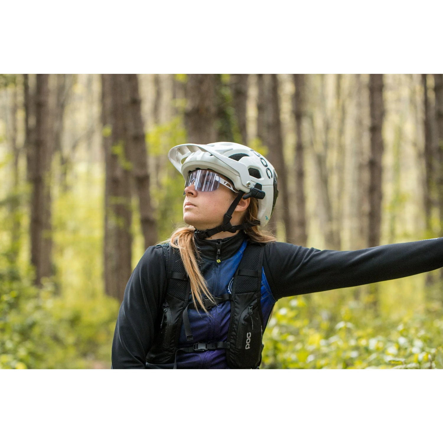 Donna con casco bianco e occhiali da sole in foresta, indossa giacca nera e gilet blu.