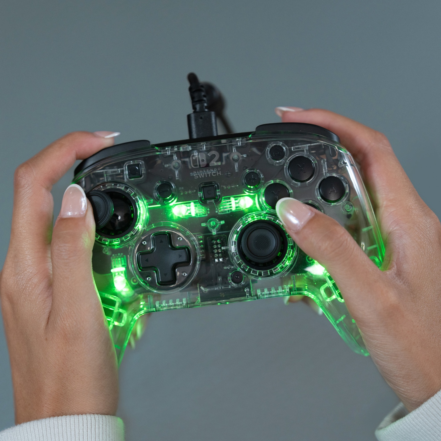 Des mains tiennent une manette transparente de Nintendo Switch éclairée en vert. Un cordon est visible.