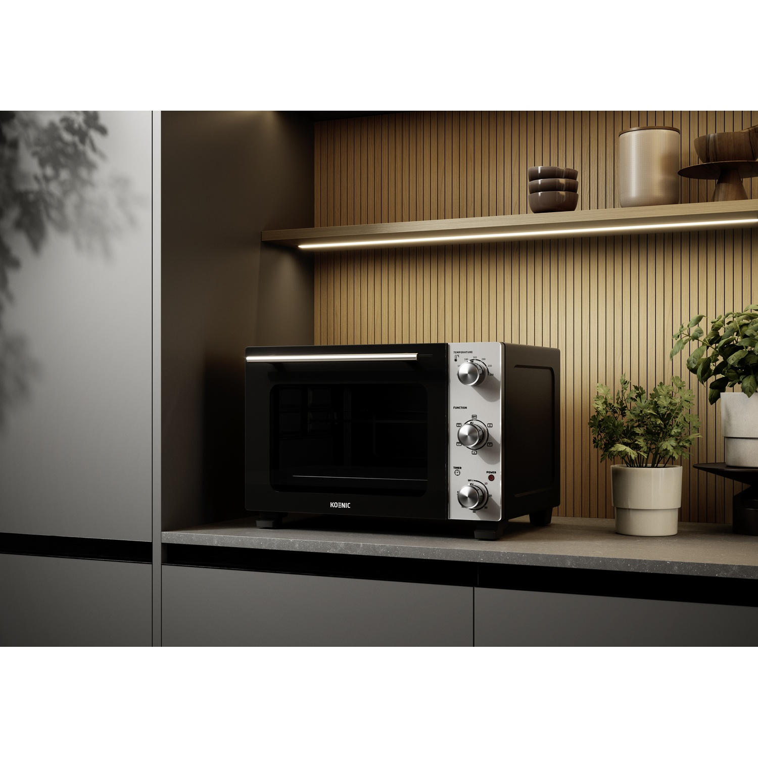 Un mini forno nero Koenic si trova su un piano di lavoro grigio con piante e decorazioni su una mensola sopra.