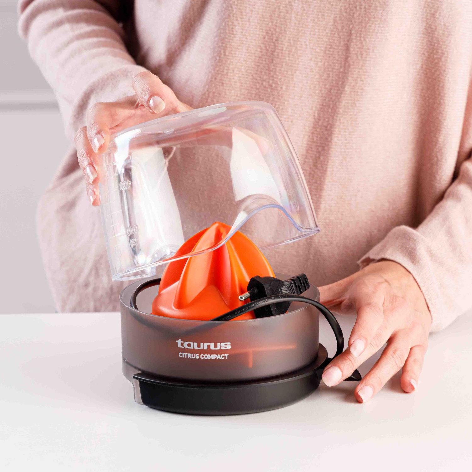 Una mujer sostiene una tapa transparente sobre un exprimidor de cítricos naranja con logo Taurus.