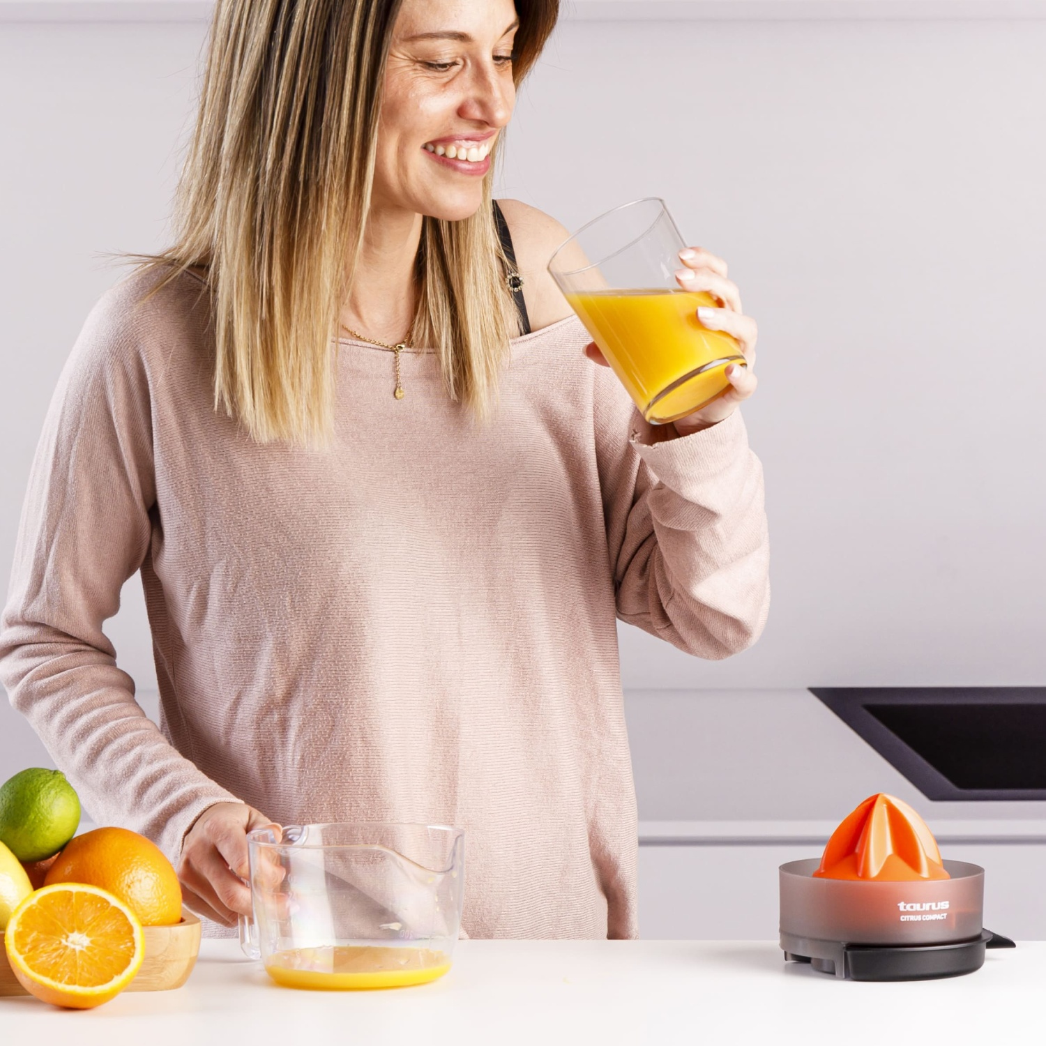 Mujer bebe jugo de naranja junto a un exprimidor y frutas.
