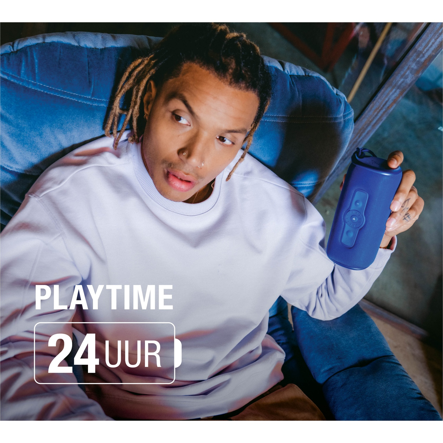 Un homme avec des dreadlocks est assis sur une chaise bleue, tenant une enceinte bleue. Texte : PLAYTIME 24 UUR.