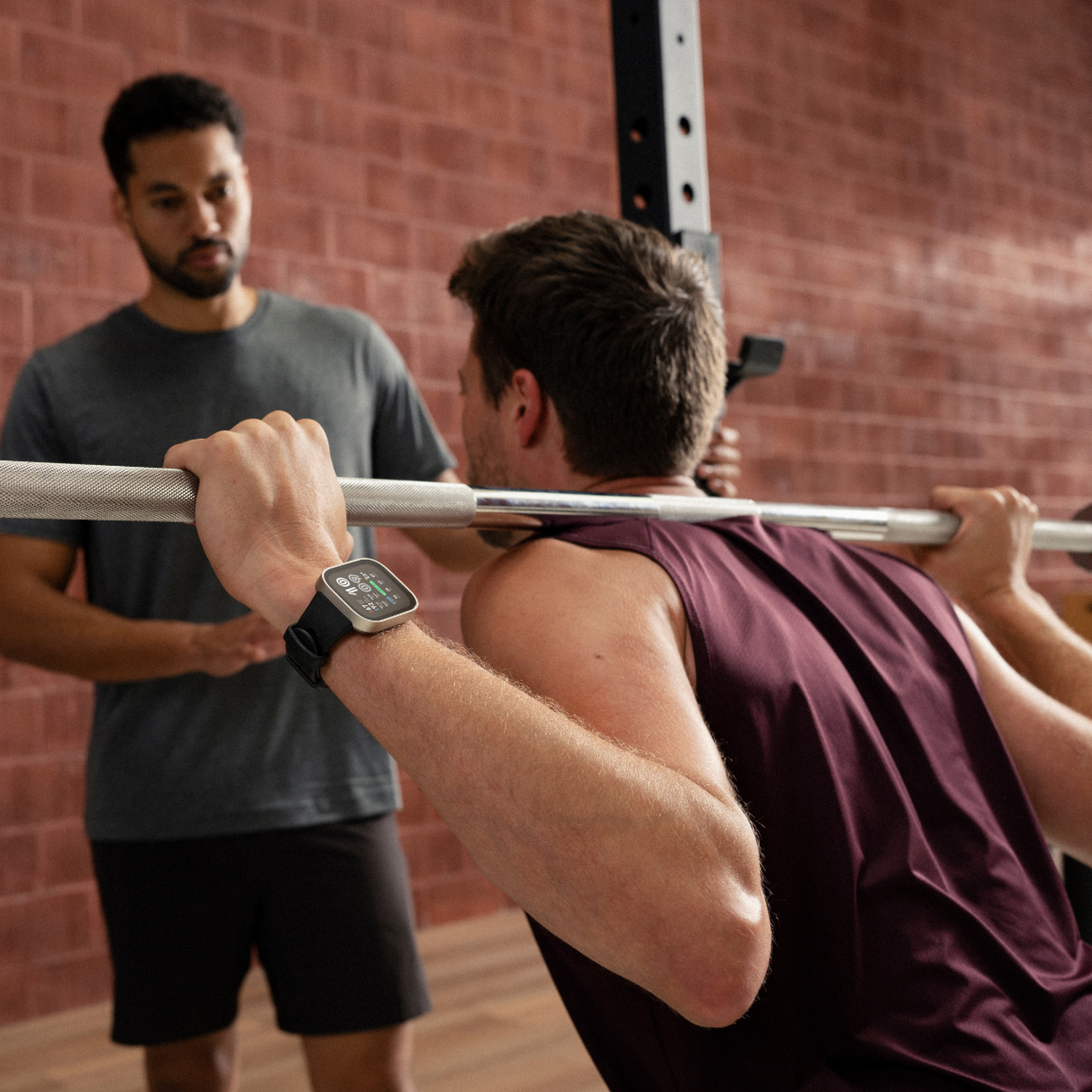 Hombre haciendo sentadillas con barra, entrenador observa. Hombre lleva un smartwatch.
