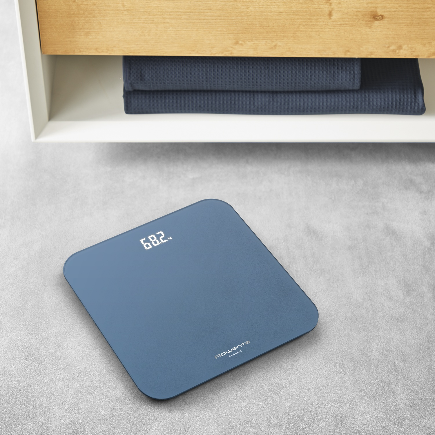 Balance numérique bleu affichant 68,2 kg, à côté de serviettes bleu foncé pliées.