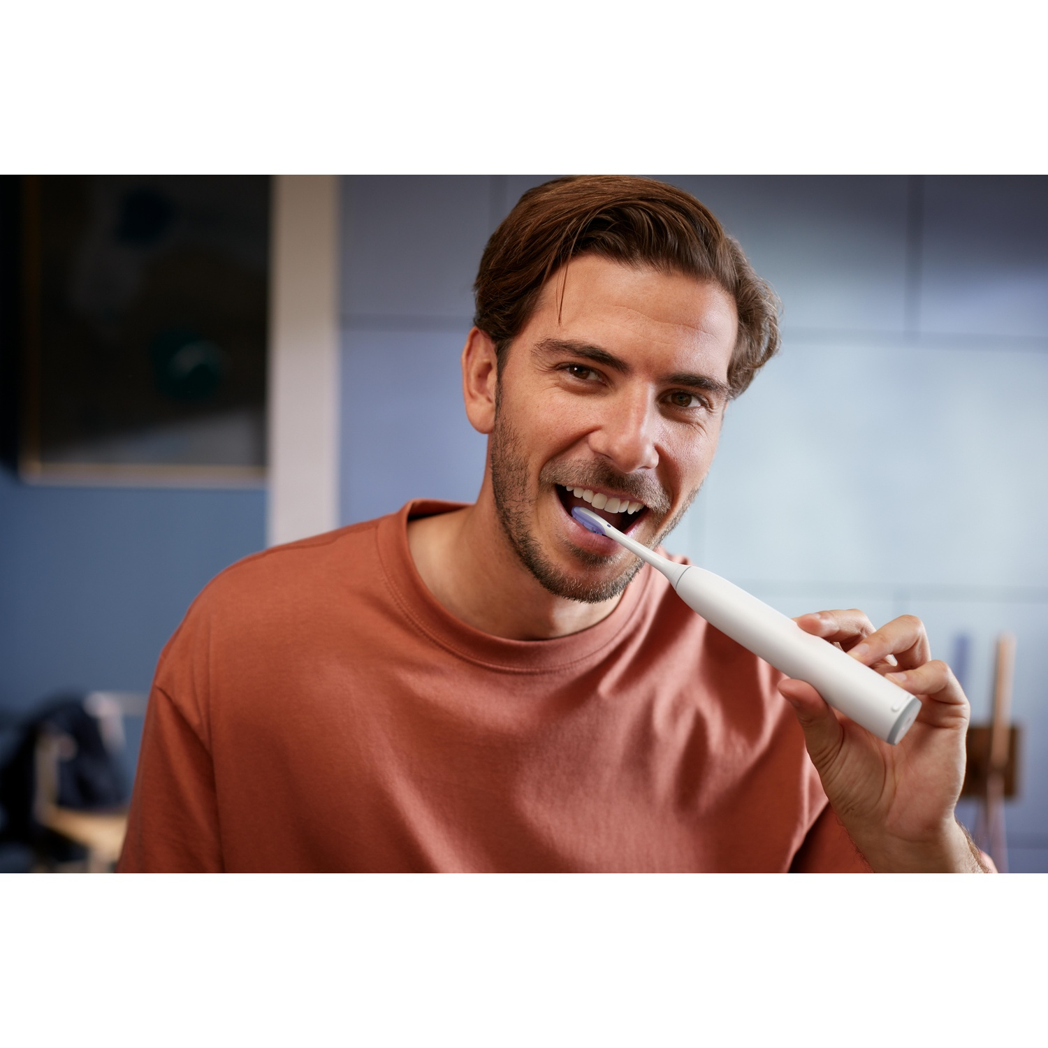Uomo con capelli castani sorride e si lava i denti con spazzolino elettrico bianco.
