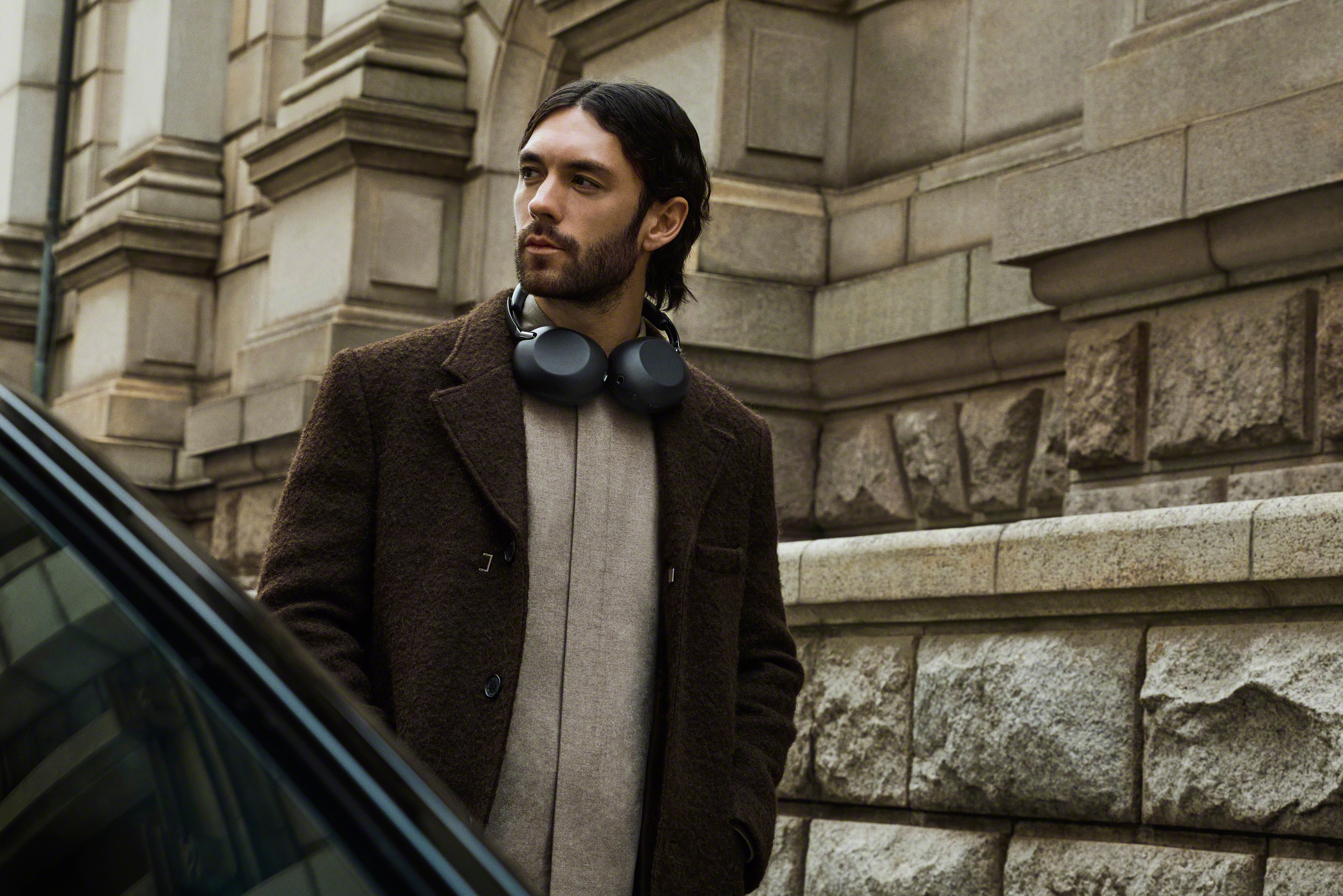 Homme avec casque autour du cou, manteau marron, devant une façade en pierre.