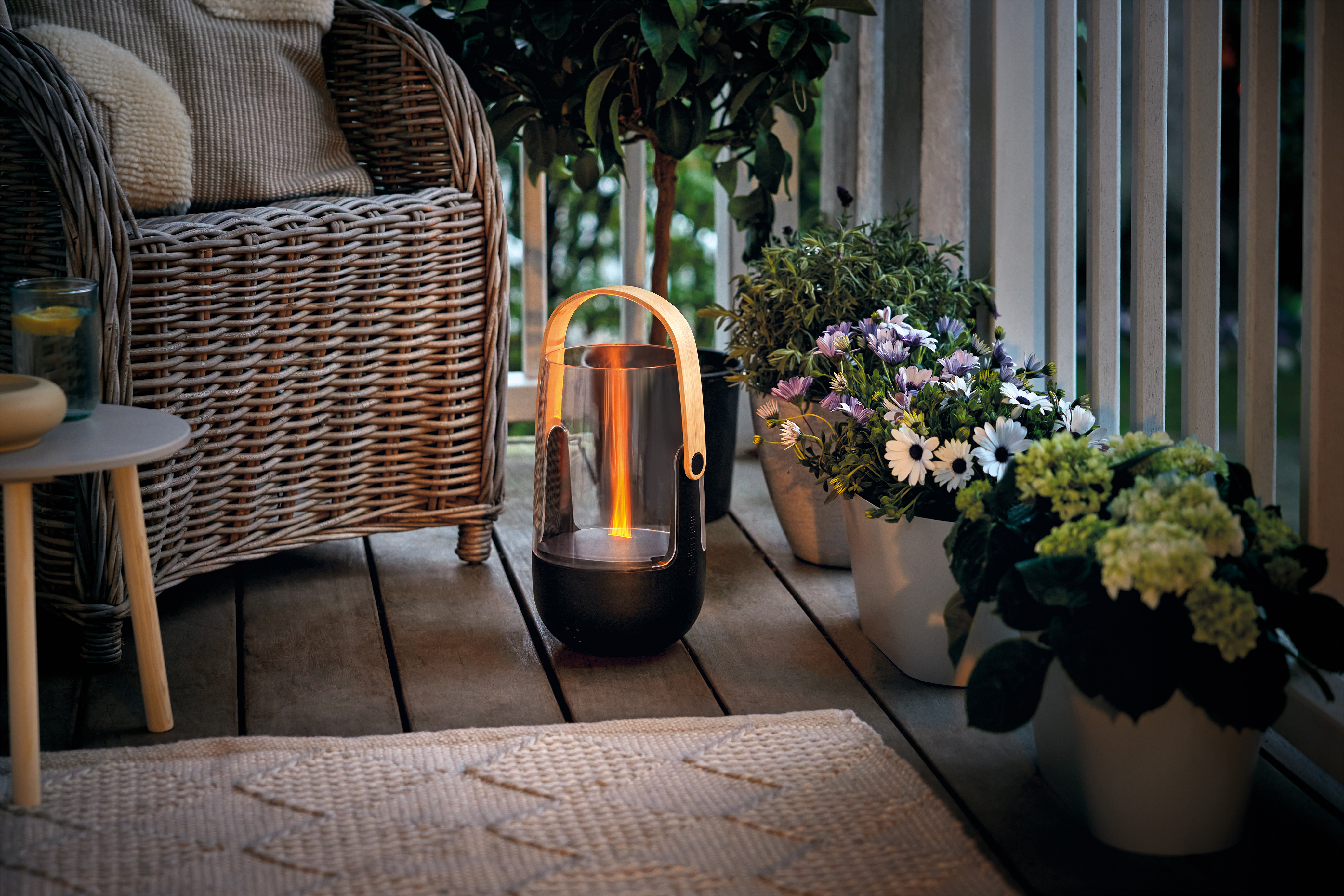 Ein Korbstuhl auf einer Holzterrasse mit einer Laterne und Blumen.