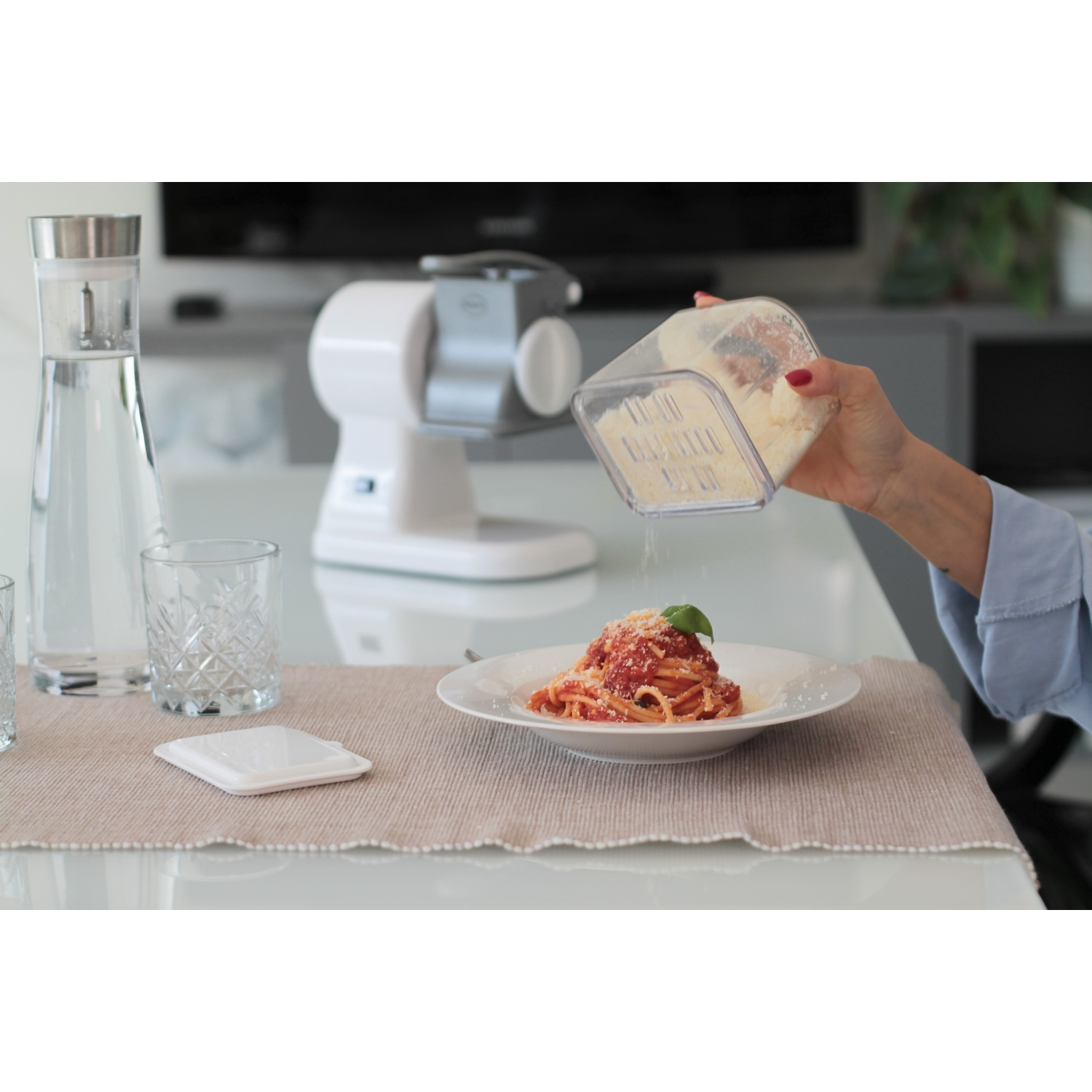 Una mano grattugia il formaggio sugli spaghetti al sugo. Una caraffa d'acqua e bicchieri sono vicini.