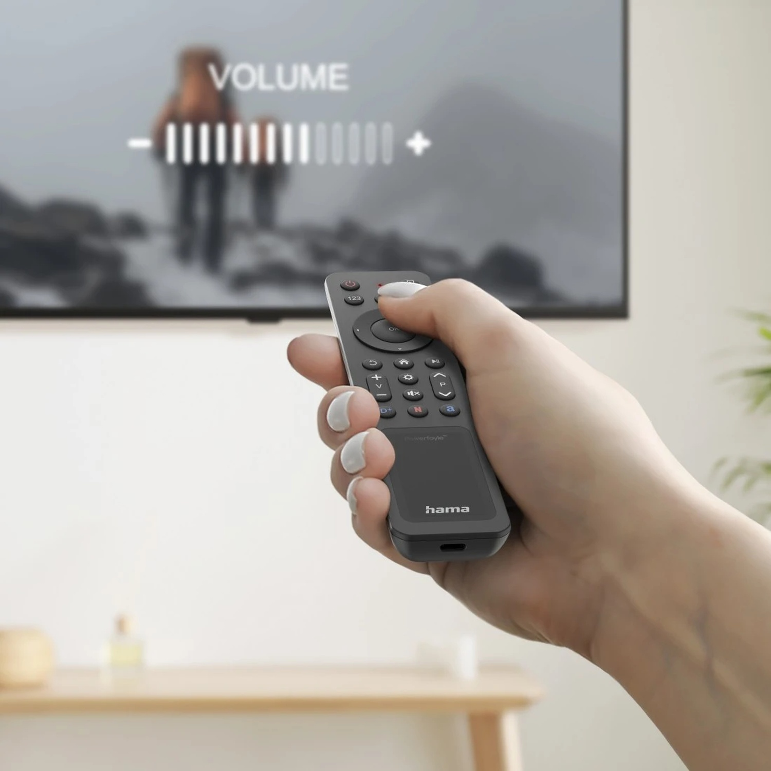 Main tenant une télécommande Hama, ajustant le volume TV. Arrière-plan flou de randonneurs.