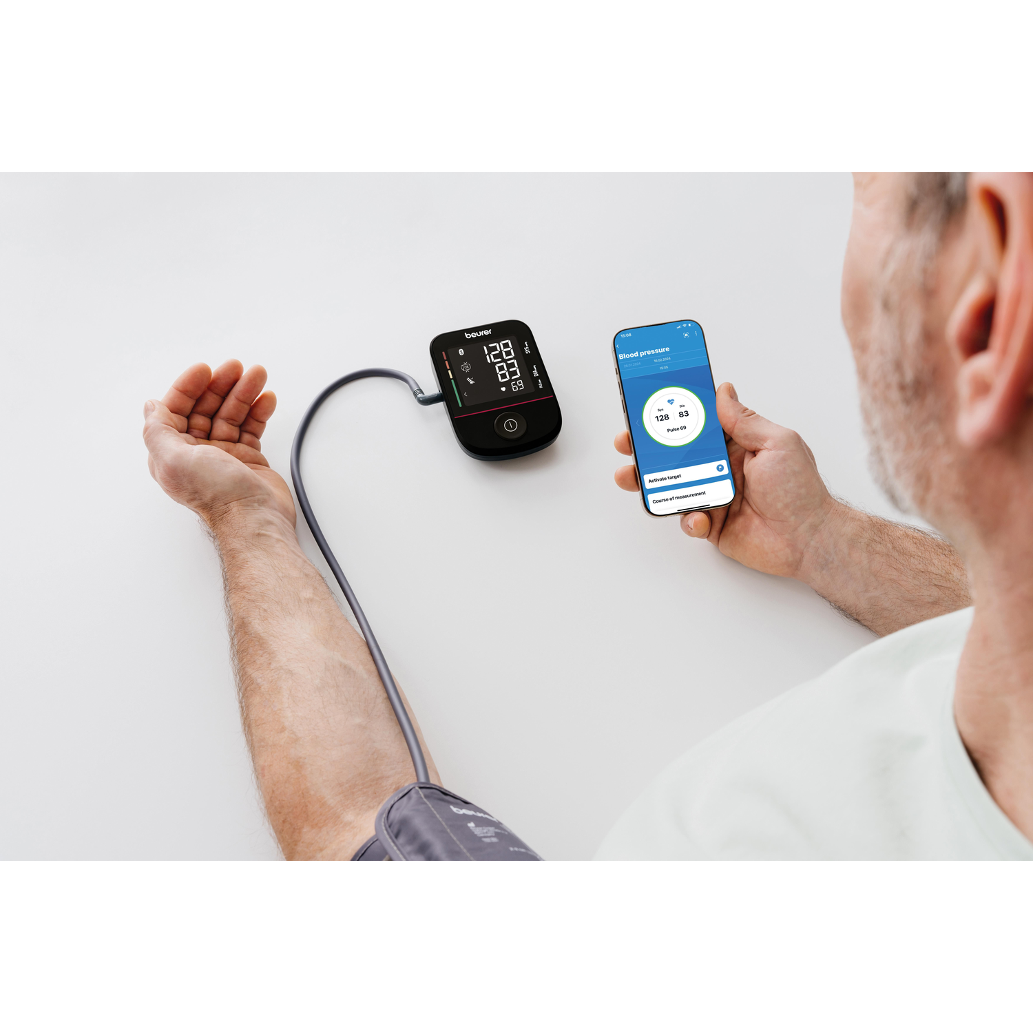Man uses blood pressure monitor connected to arm, displaying results on device and phone.