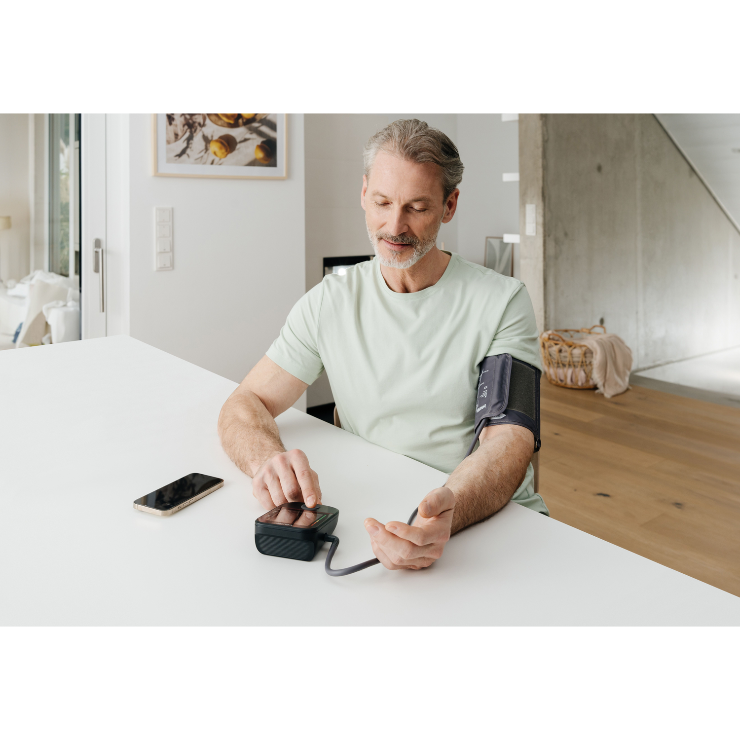 Man measures blood pressure at home. A phone and a monitor are on the table.