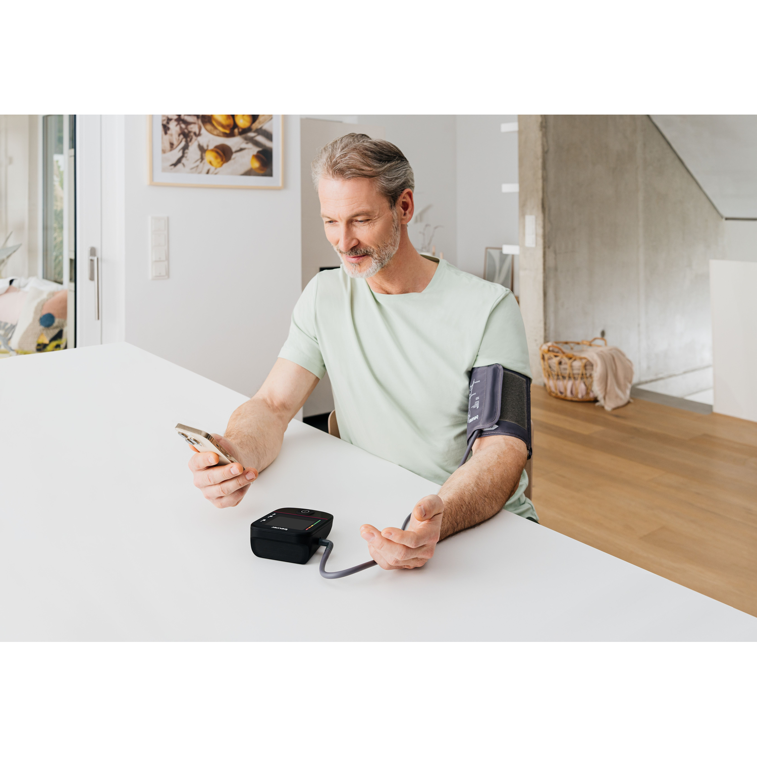Man with arm cuff and phone sits at a table, looking at device.