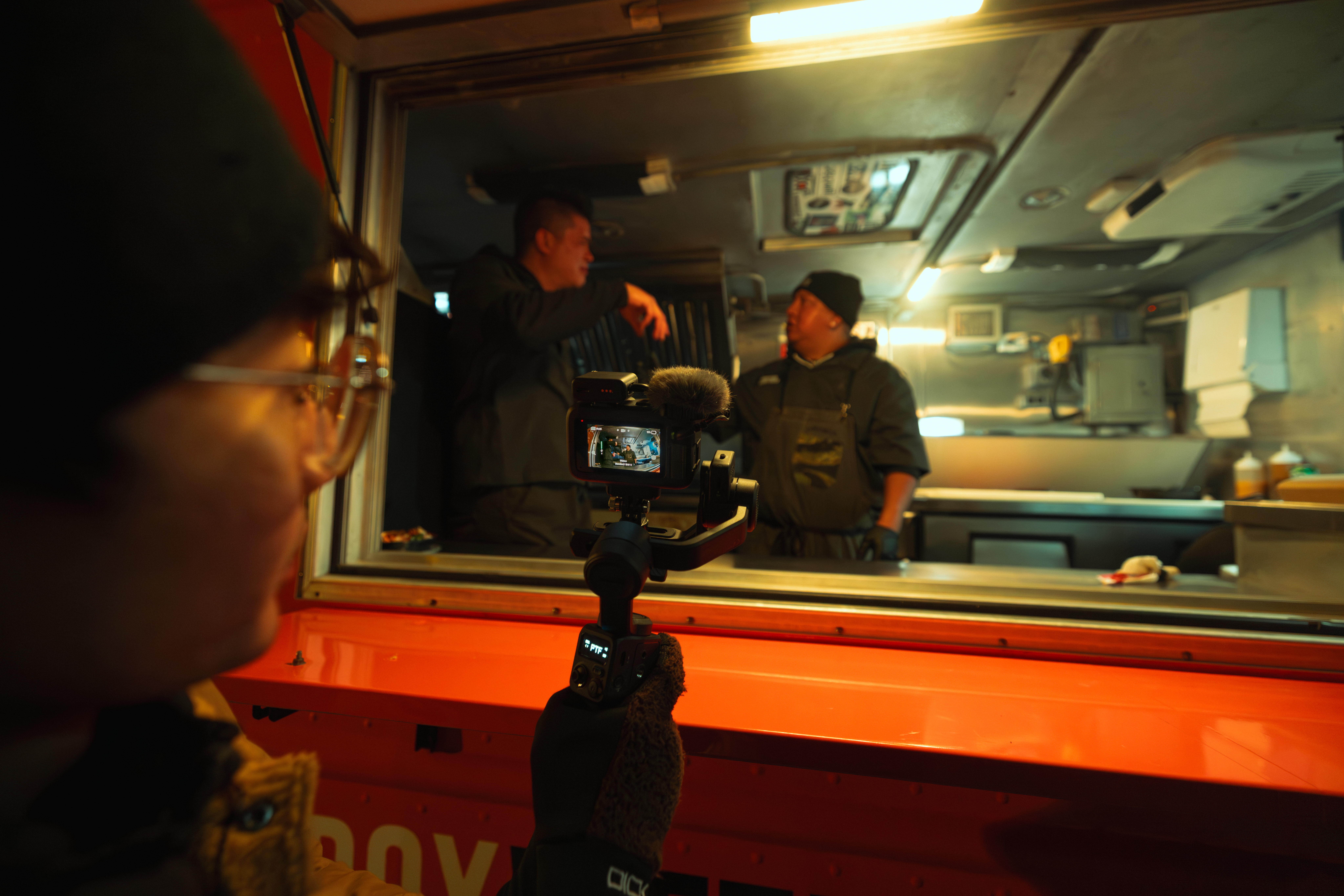 Osoba filmuje dwóch mężczyzn pracujących za ladą food trucka.