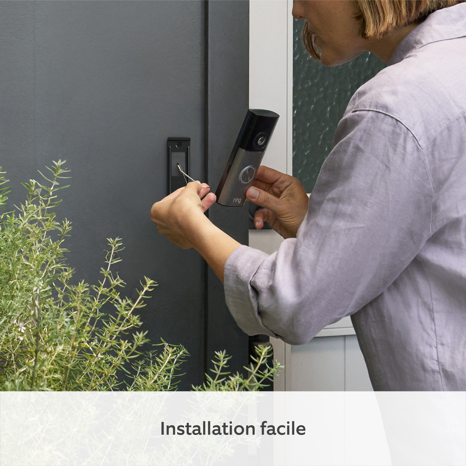 Une personne installe une sonnette vidéo sur une porte grise près de plantes vertes.